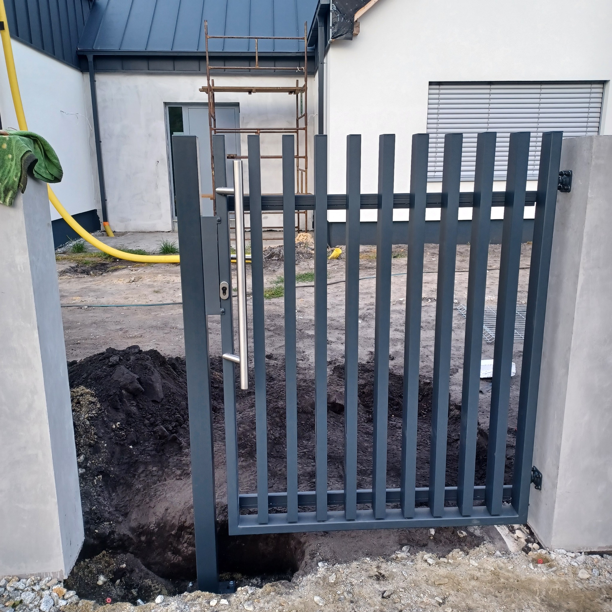 Szara furtka z pionowymi szczeblami i srebrną klamką, osadzona między betonowymi słupkami na placu budowy, z widoczną stertą ziemi i żółtym wężem w tle.