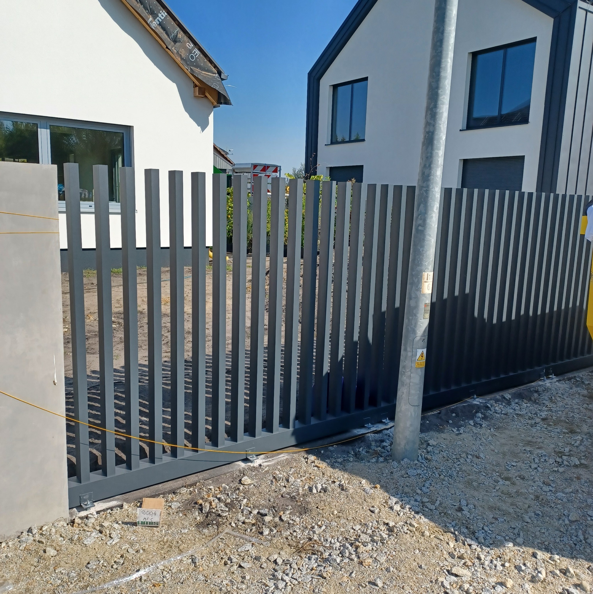 Nowoczesna, przesuwna brama wjazdowa z pionowych, metalowych profili w kolorze grafitowym, zamontowana na posesji z dwoma budynkami o minimalistycznej architekturze, widoczna skrzynka z akcesoriami...