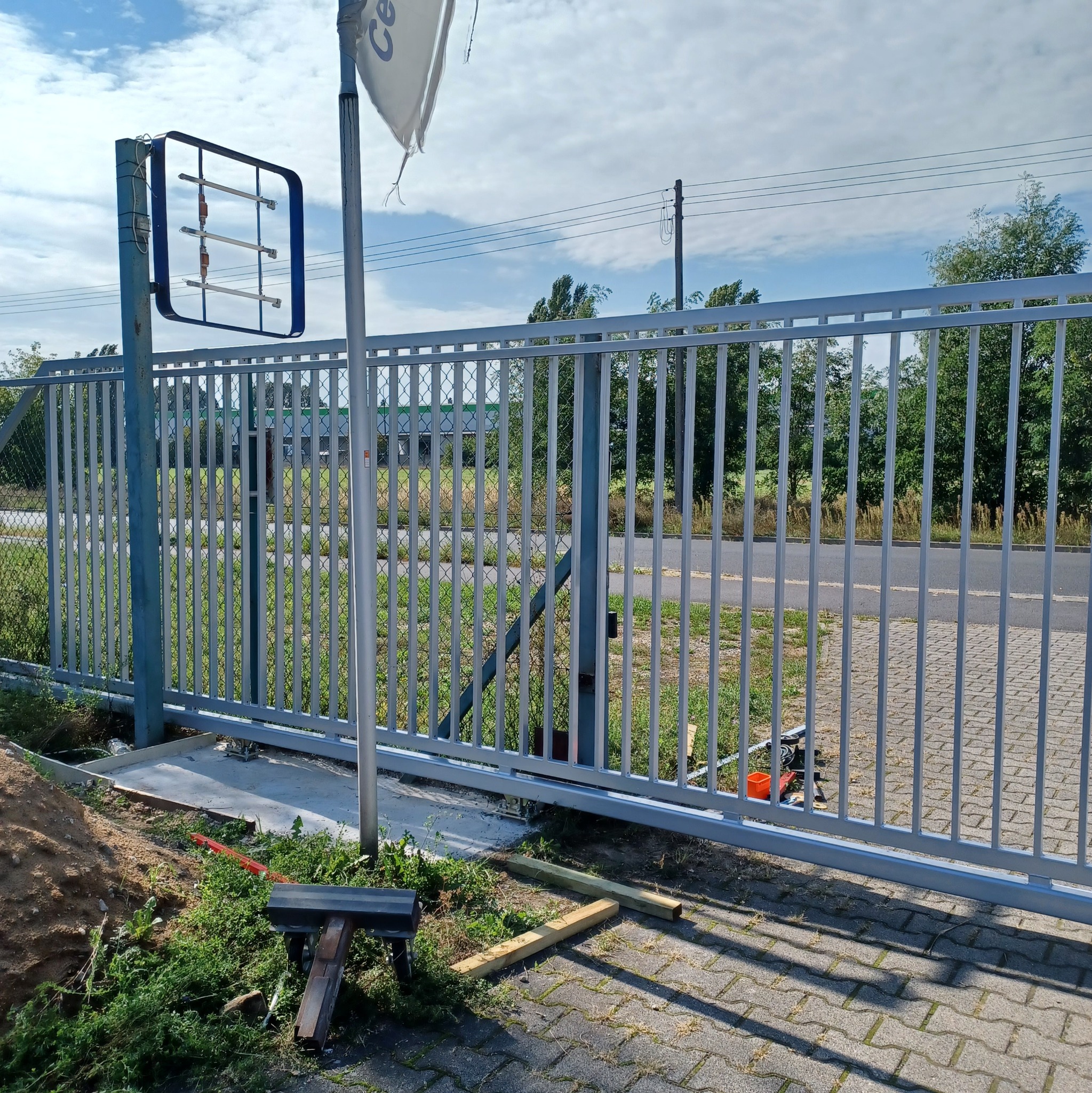 Szara, przesuwna brama wjazdowa z pionowymi prętami w trakcie montażu, widoczne narzędzia i materiały budowlane na kostce brukowej.