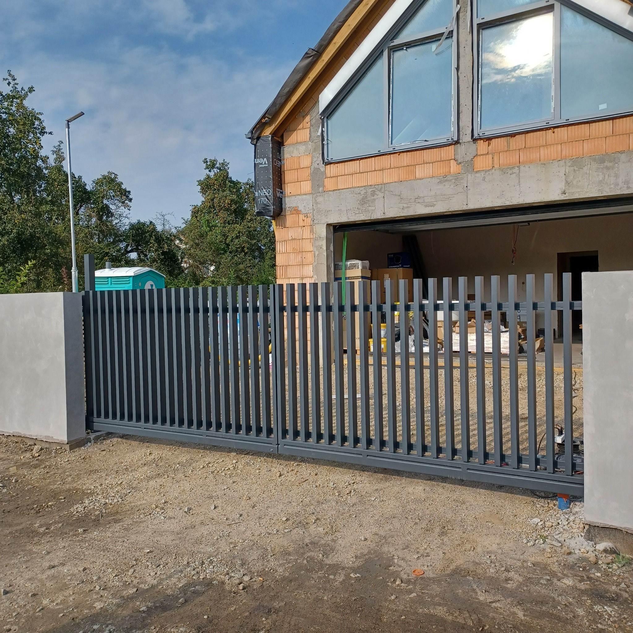 Nowoczesna, przesuwna, metalowa brama w kolorze grafitowym, z pionowymi szczeblami, zamontowana przed surowym budynkiem w trakcie budowy, z widocznymi pustakami i oknami.