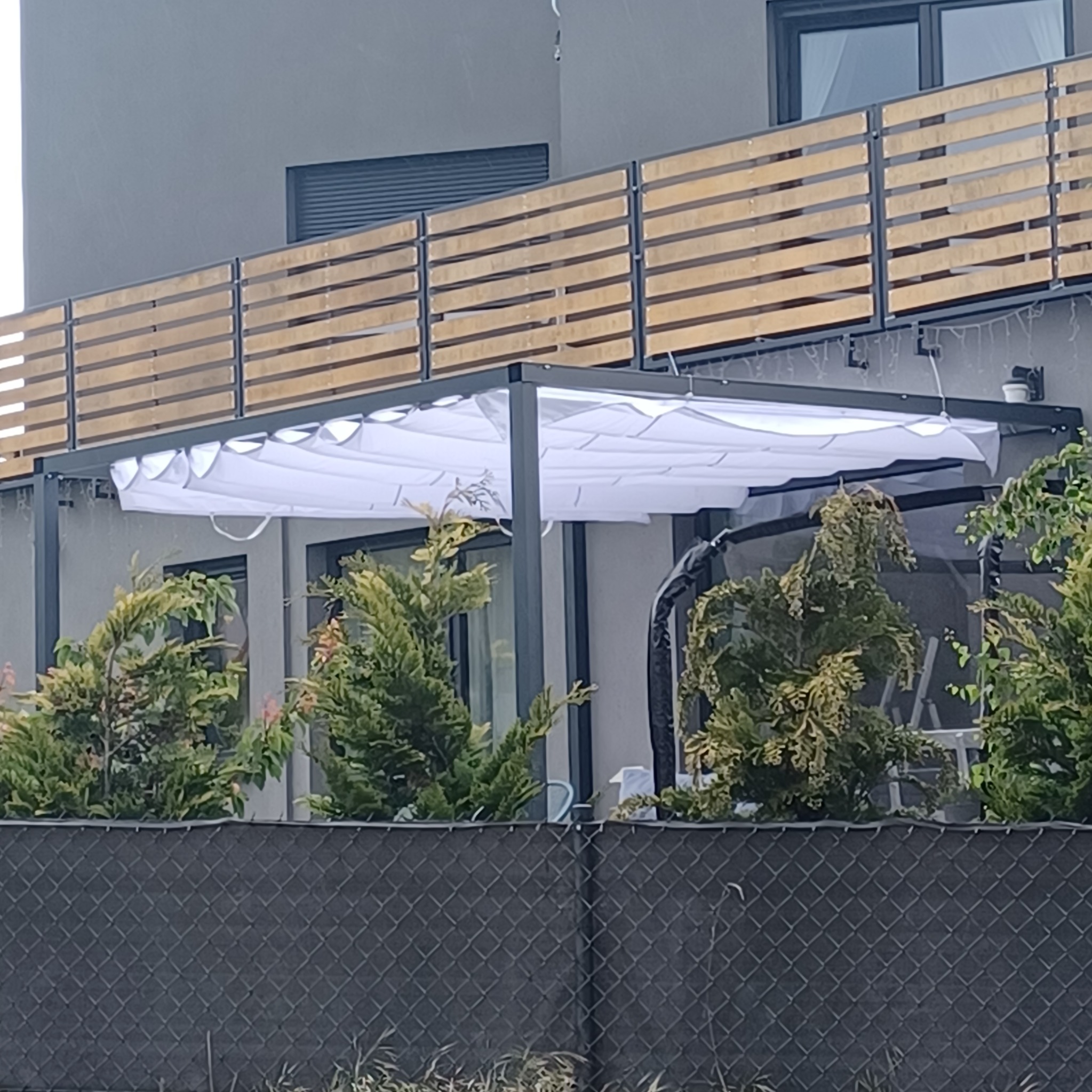 Zadaszenie tarasu z białym, materiałowym dachem harmonijkowym, widoczne zza siatkowego ogrodzenia i iglaków, z drewnianą balustradą balkonu powyżej.