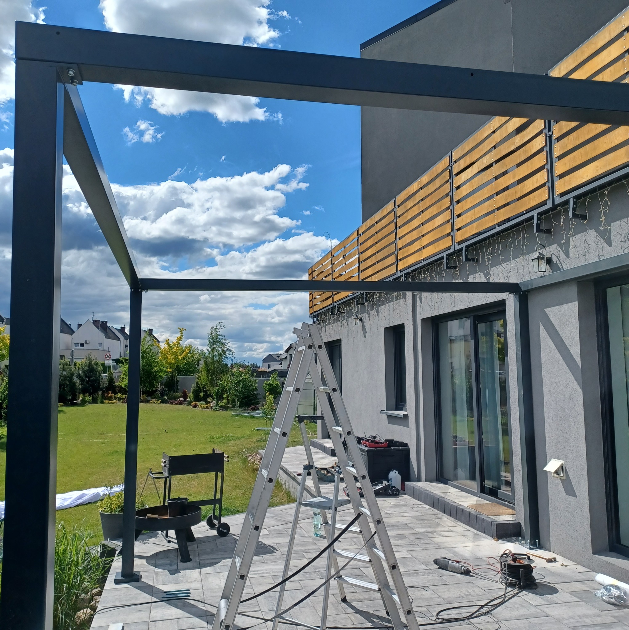 Montaż metalowej konstrukcji z drabiny na tarasie, widoczne narzędzia i elementy mocujące, tło to dom z drewnianą balustradą i ogród.