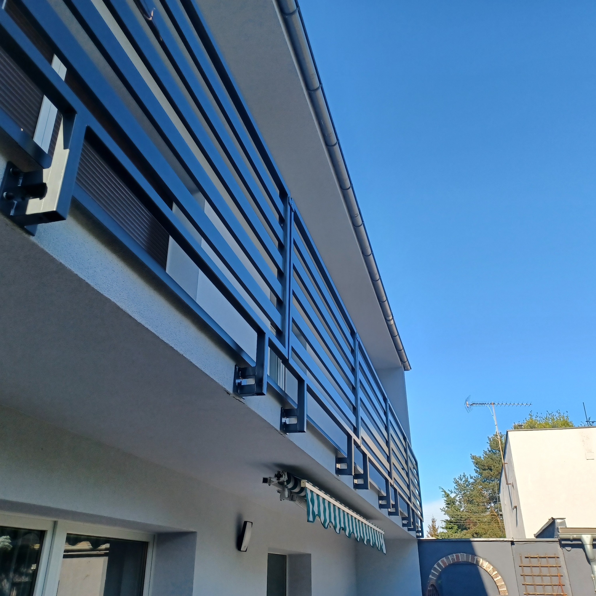 Nowoczesna balustrada balkonowa z czarnych, poziomych profili, zamontowana do jasnej elewacji budynku, widok z dołu na błękitne niebo.