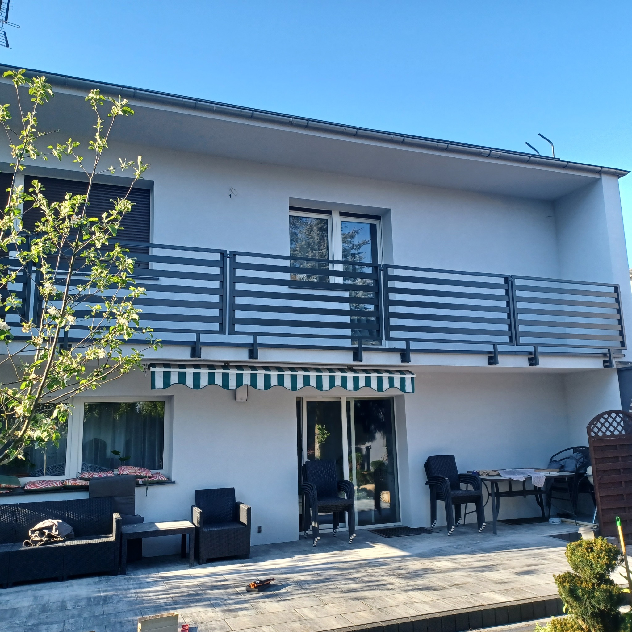 Nowoczesny dom z szarą elewacją, balkonem z poziomą balustradą ze stali nierdzewnej i tarasem z meblami ogrodowymi, oświetlony porannym słońcem.