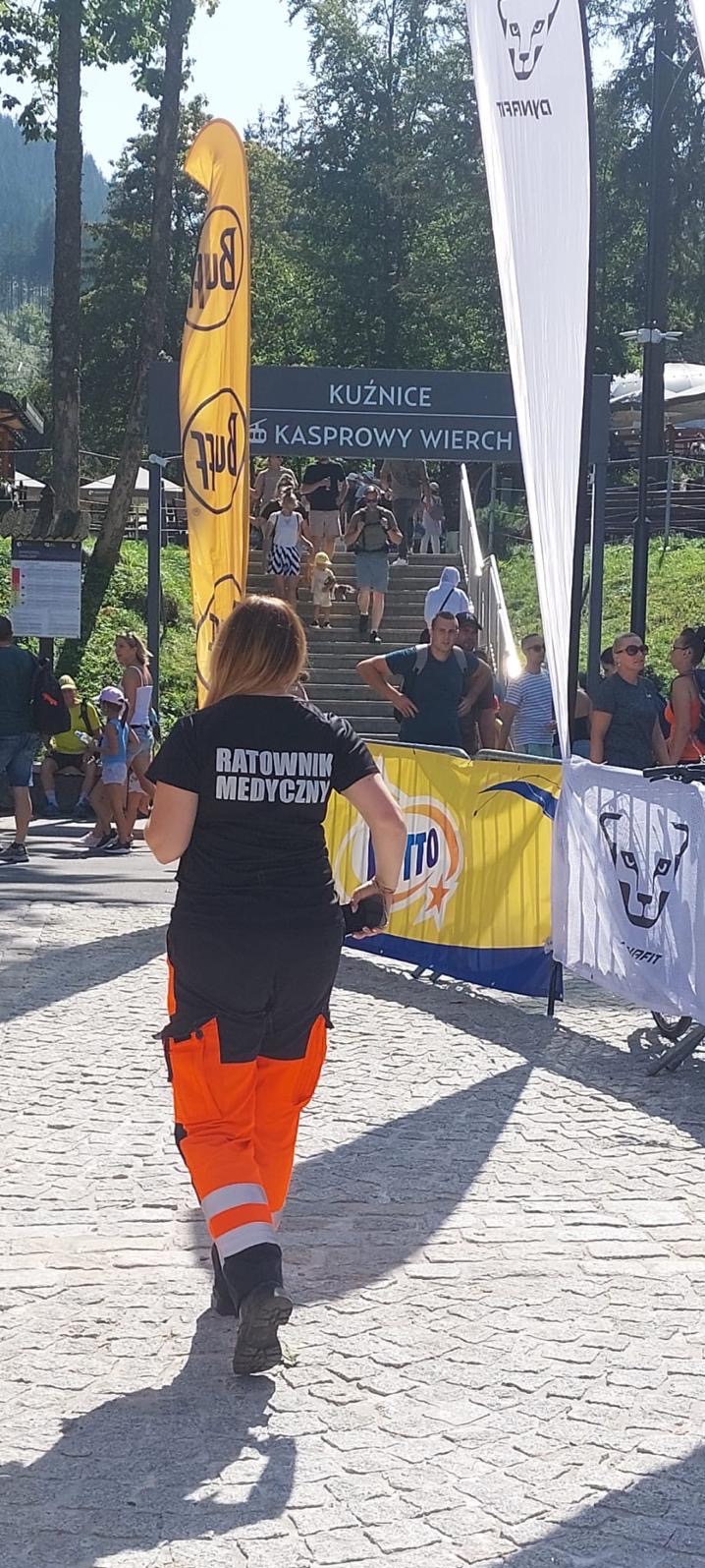Ratownik medyczny w czarnym uniformie z pomarańczowymi spodniami idzie po brukowanej drodze w kierunku schodów prowadzących do Kasprowego Wierchu, otoczony przez flagi reklamowe i przechodzących...