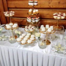 Elegancki stół z deserami: babeczki z owocami, mini torty w kształcie serc i lizaki, ustawione na złotych paterach i szklanych naczyniach, udekorowany białymi różami.