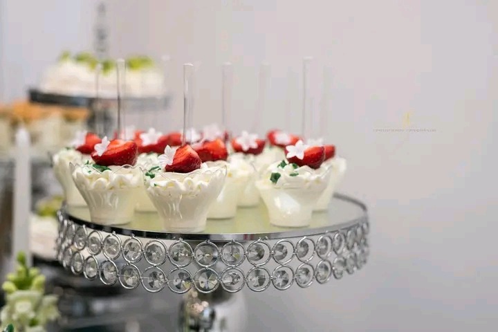 Deserowe babeczki z kremem i truskawkami na eleganckiej, zdobionej paterze. Białe tło, delikatne oświetlenie, widoczne inne desery w tle.