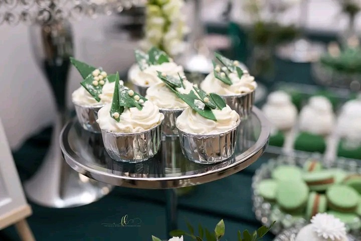 Eleganckie babeczki z białym kremem i zielonymi dekoracjami na srebrnej paterze, część zastawy i słodkości w tle, stonowana kolorystyka.
