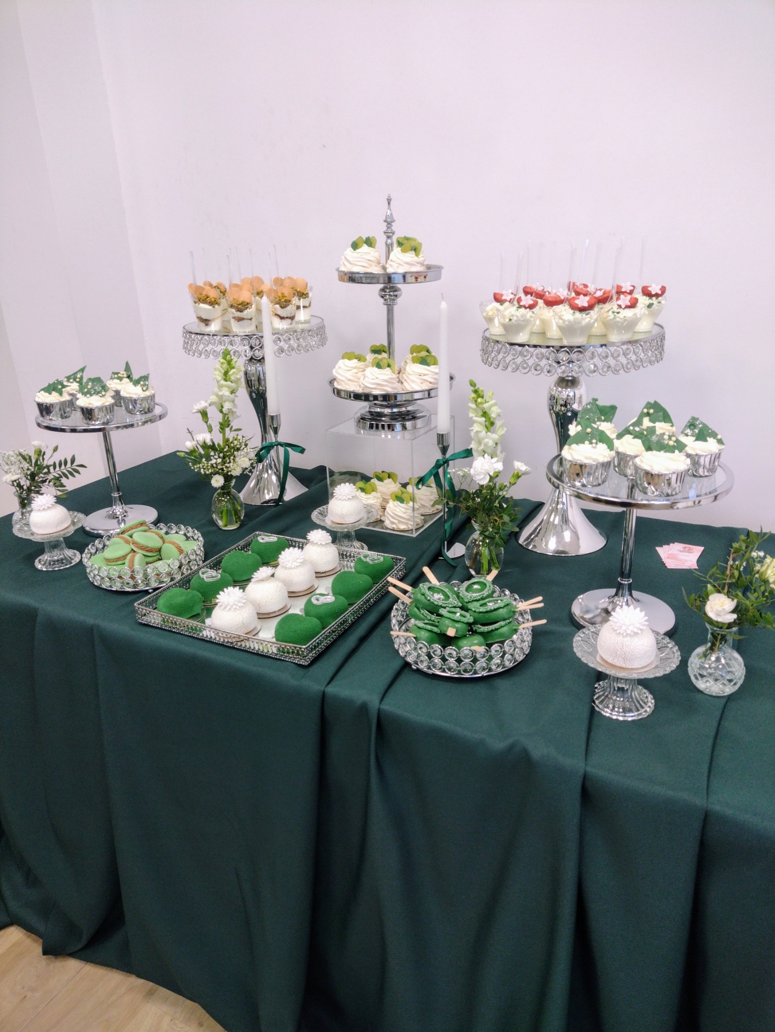 Ekskluzywny stół z deserami: zielone makaroniki, babeczki i mini torty na srebrnych paterach. Dekoracje kwiatowe i świece dodają elegancji. Idealne na przyjęcie.