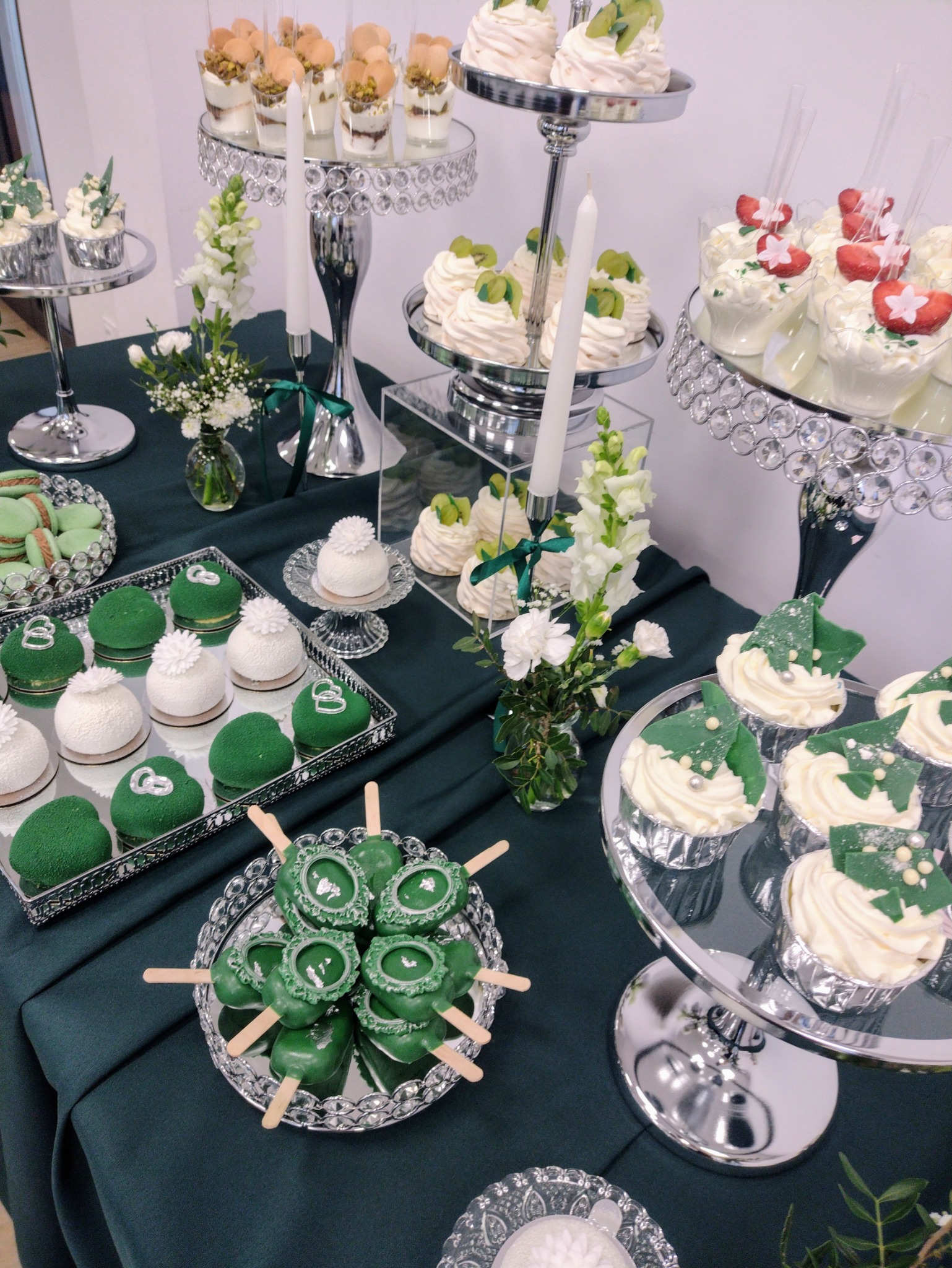 Elegancki stół cukierniczy z zielonymi dekoracjami: babeczki, cake pops, mini desery, makaroniki i kwiaty na srebrnych paterach. Idealne na uroczystości.