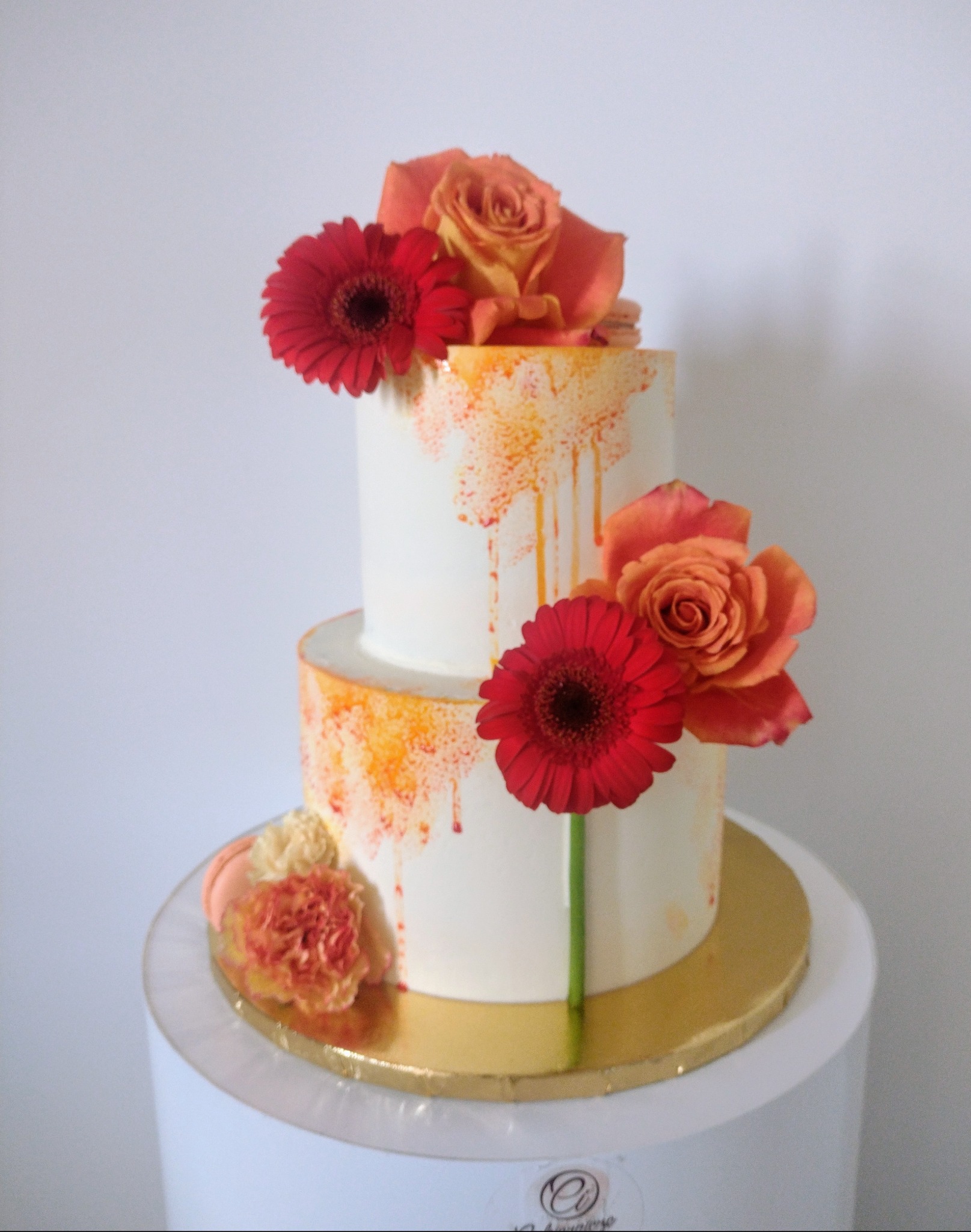 Dwupoziomowy tort weselny z białym kremem, pomarańczowymi akcentami i dekoracją z róż oraz gerber na złotym podkładzie.