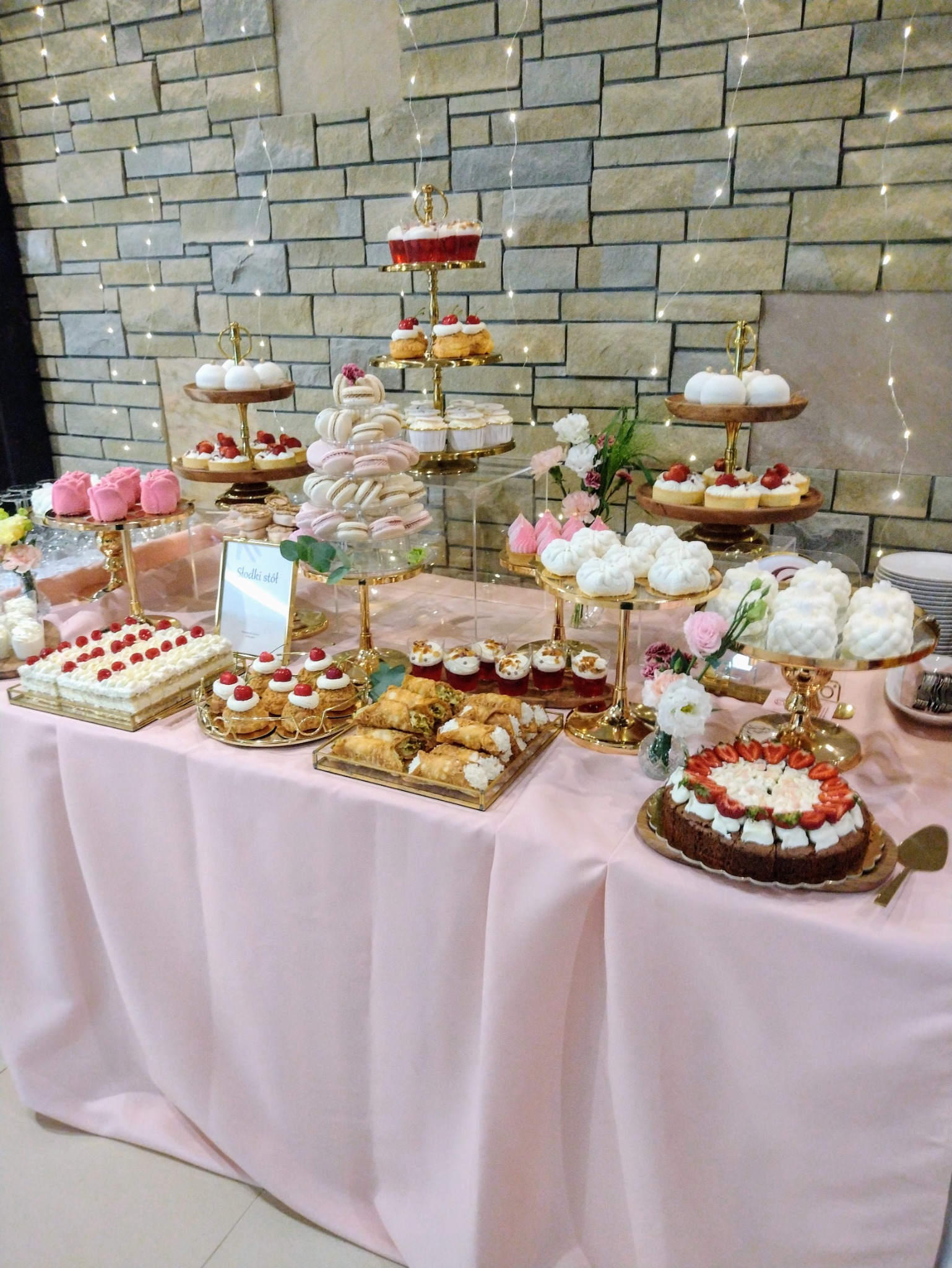 Słodki stół z tortami, babeczkami i makaronikami na różowym obrusie, dekorowany lampkami, ustawiony na tle kamiennej ściany.