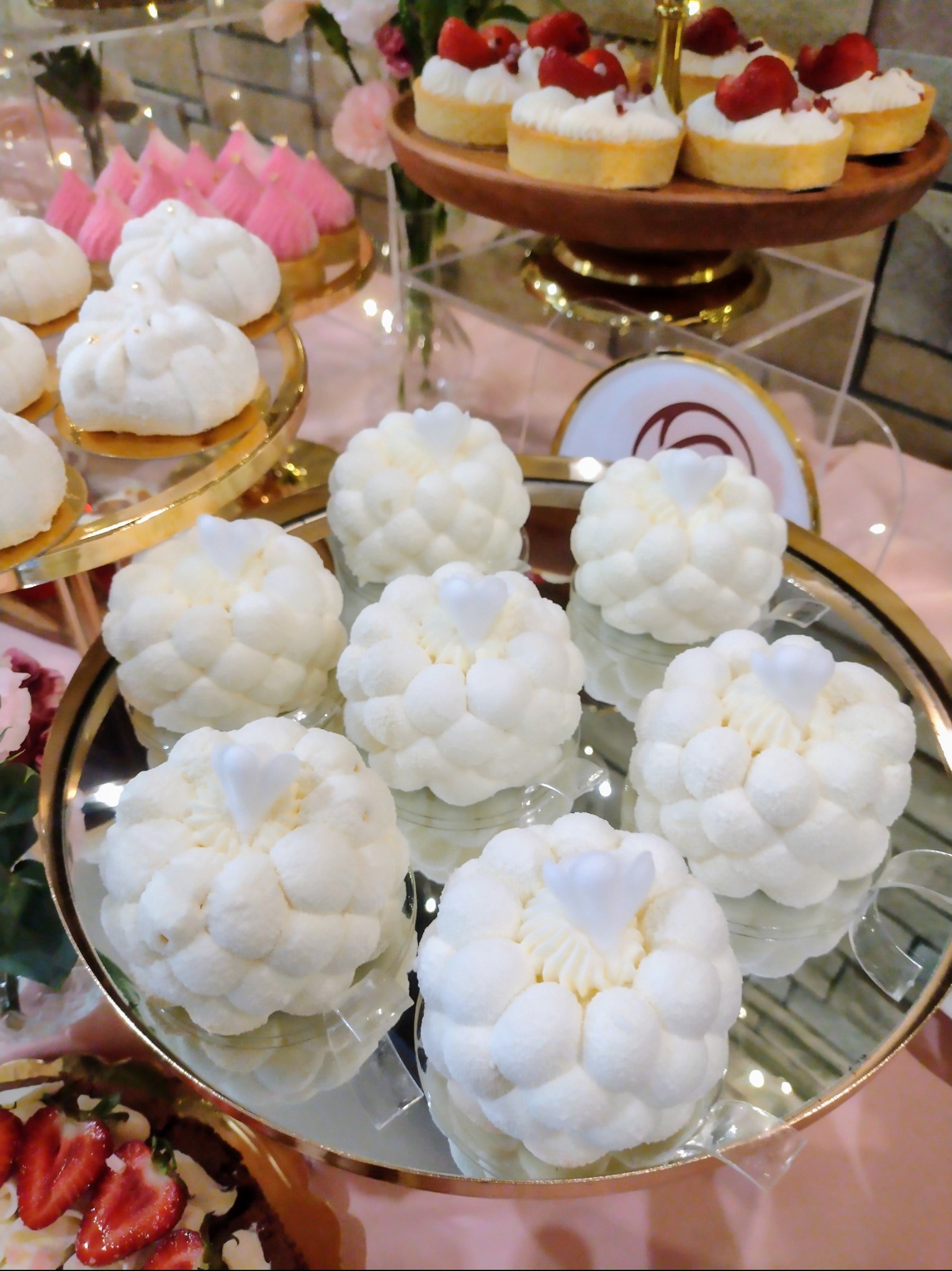 Eleganckie, białe mini-desery z serduszkami na lustrzanej tacy, część wystawnego stołu z tartaletkami i różowymi słodkościami w tle. Idealne na przyjęcie.