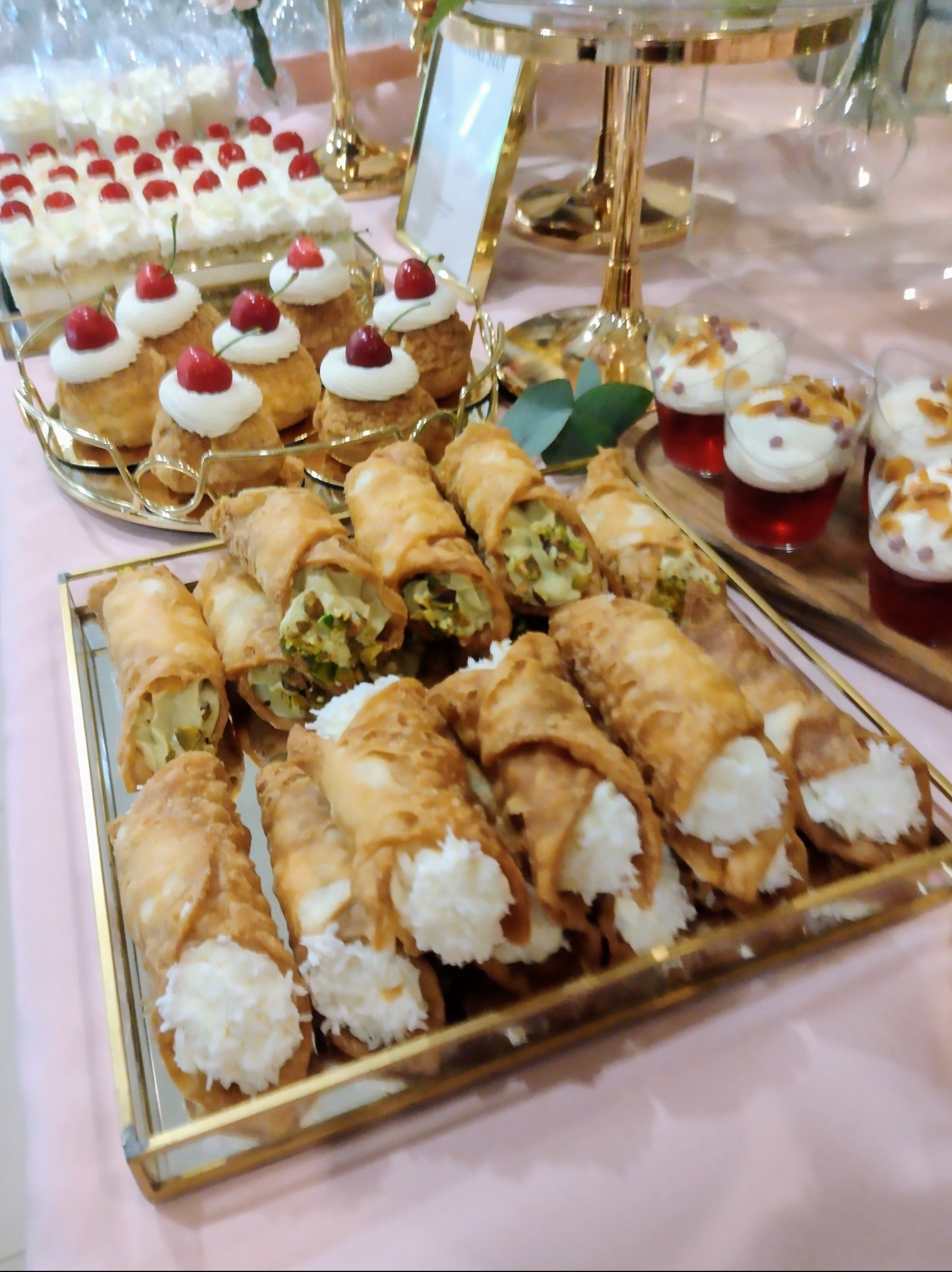 Elegancki stół z deserami: cannoli z kremem kokosowym i pistacjowym, babeczki z wiśniami i galaretki z bitą śmietaną, idealne na przyjęcie.