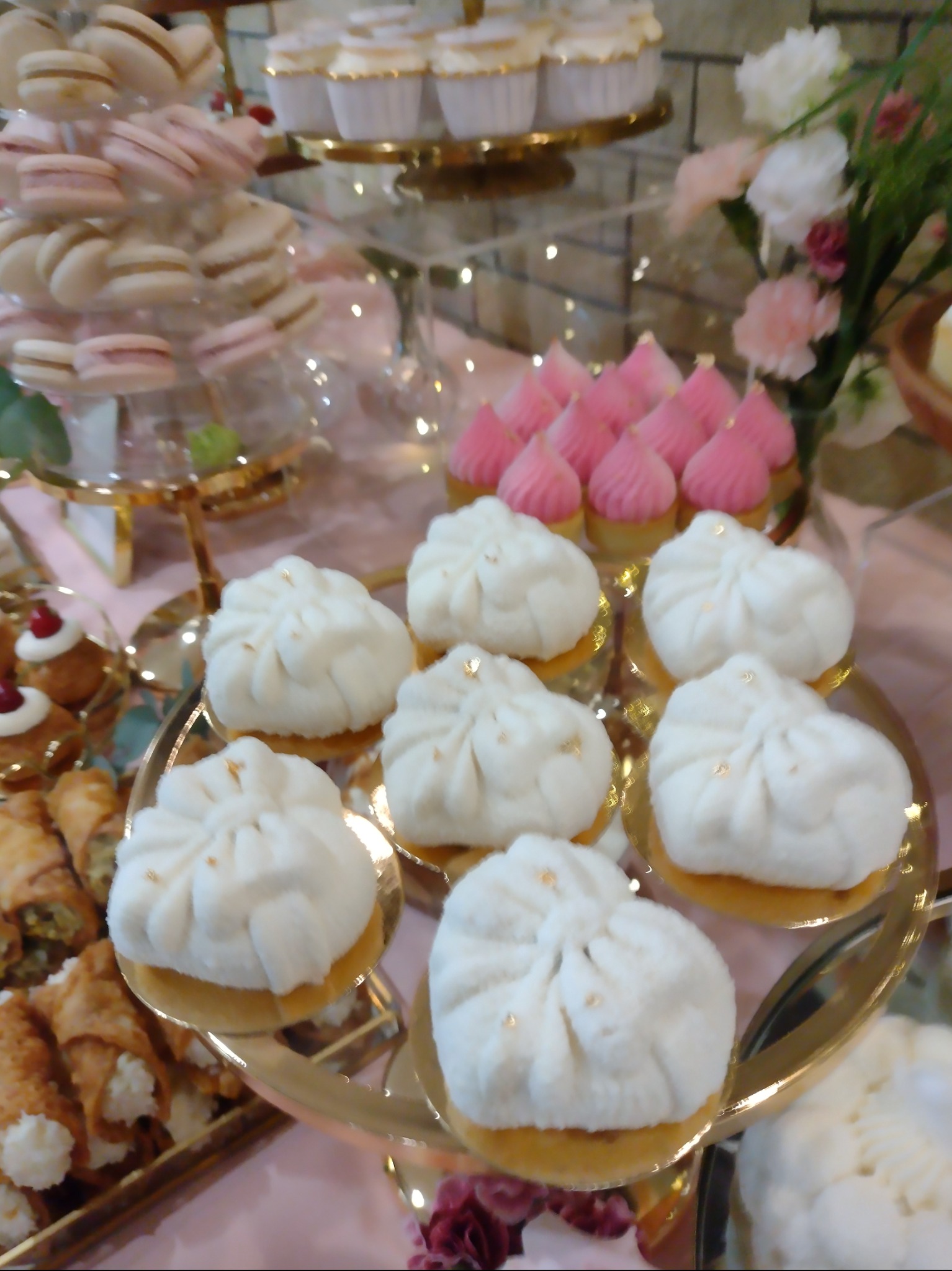 Słodki stół z różnorodnymi deserami: białe babeczki z dekoracją, makaroniki na stojaku, różowe mini-babeczki i włoskie rurki z kremem na złotych paterach.