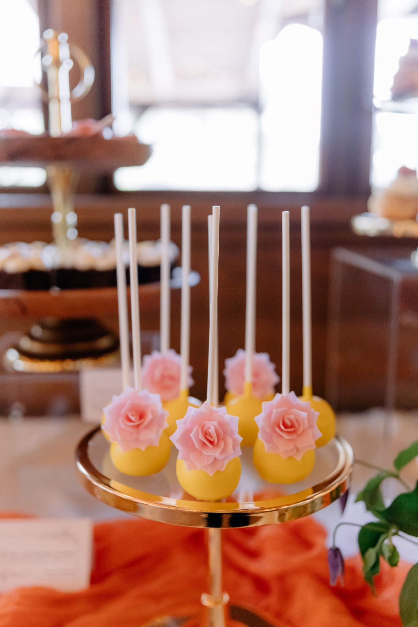 Żółte cake pops z różowymi różami na złotym stojaku, część zastawionego stołu z deserami w tle. Słodkości na przyjęcie, elegancki catering.