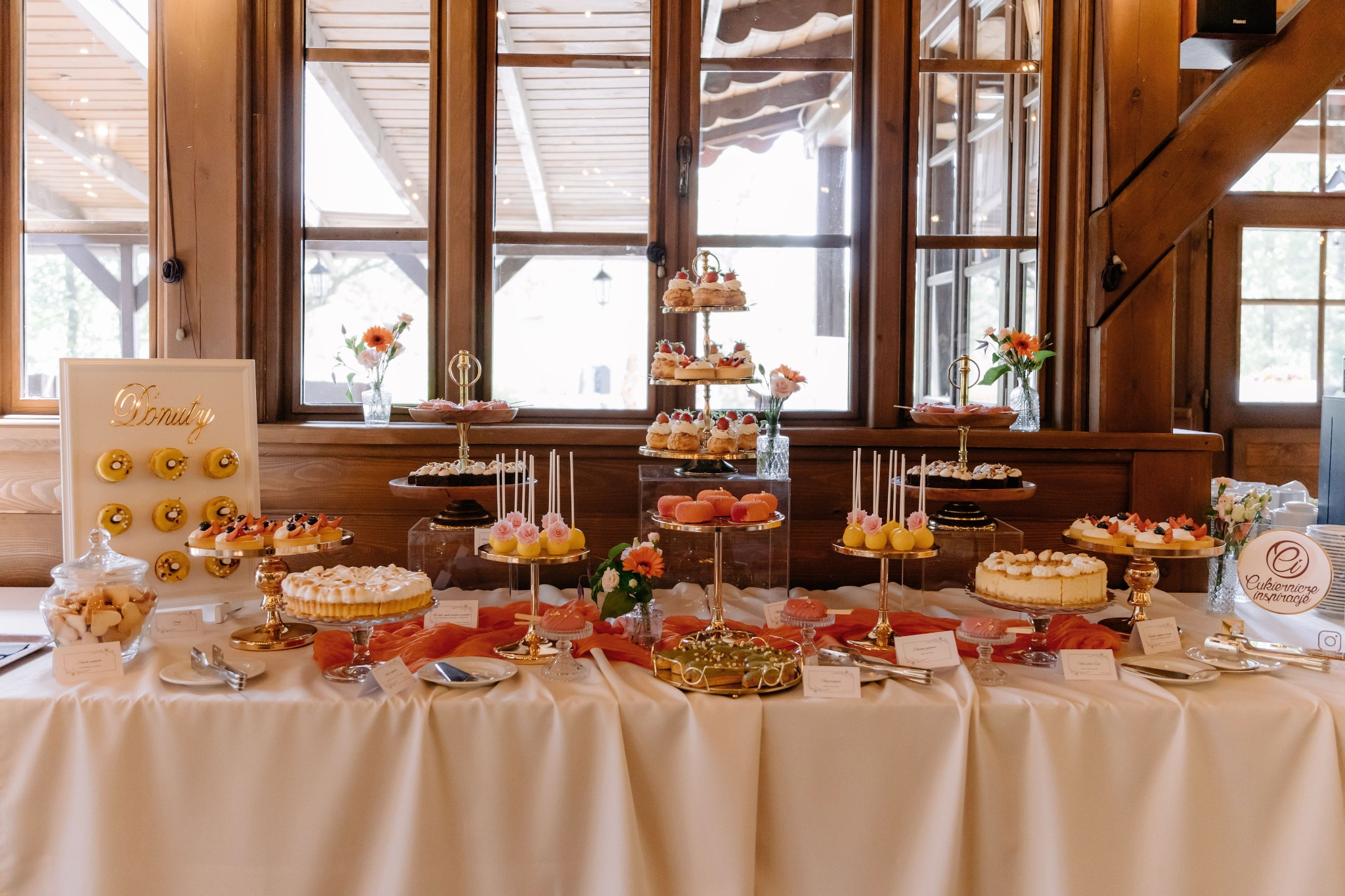 Słodki stół z ciastami, tortami i deserami na złotych paterach, dekorowany pomarańczową tkaniną, na tle drewnianej ściany z oknami.