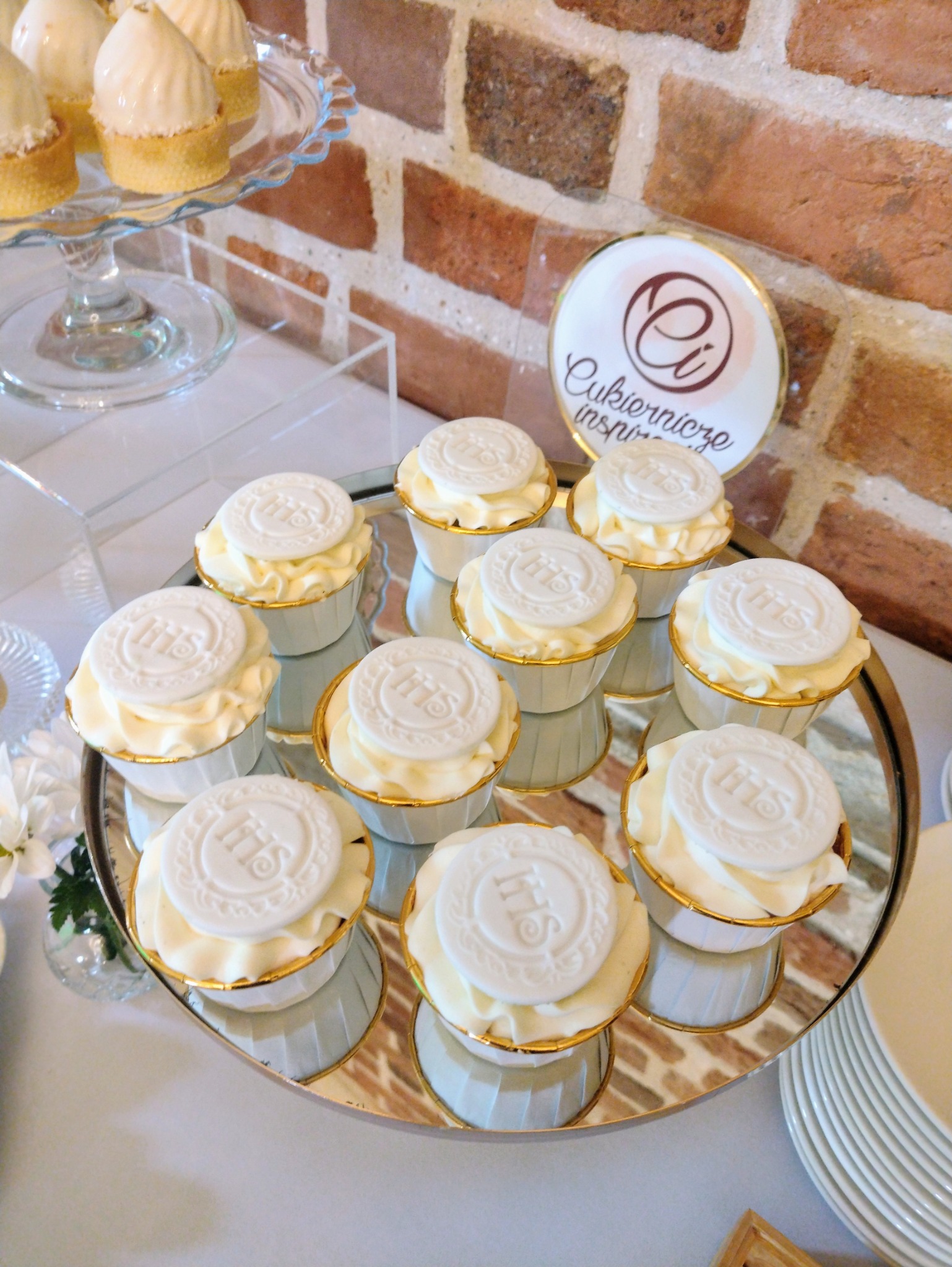 Eleganckie babeczki z białym kremem i dekoracją IHS na srebrnej tacy, ustawione na tle ceglanej ściany i logo cukierni, tworzą wyrafinowaną kompozycję.
