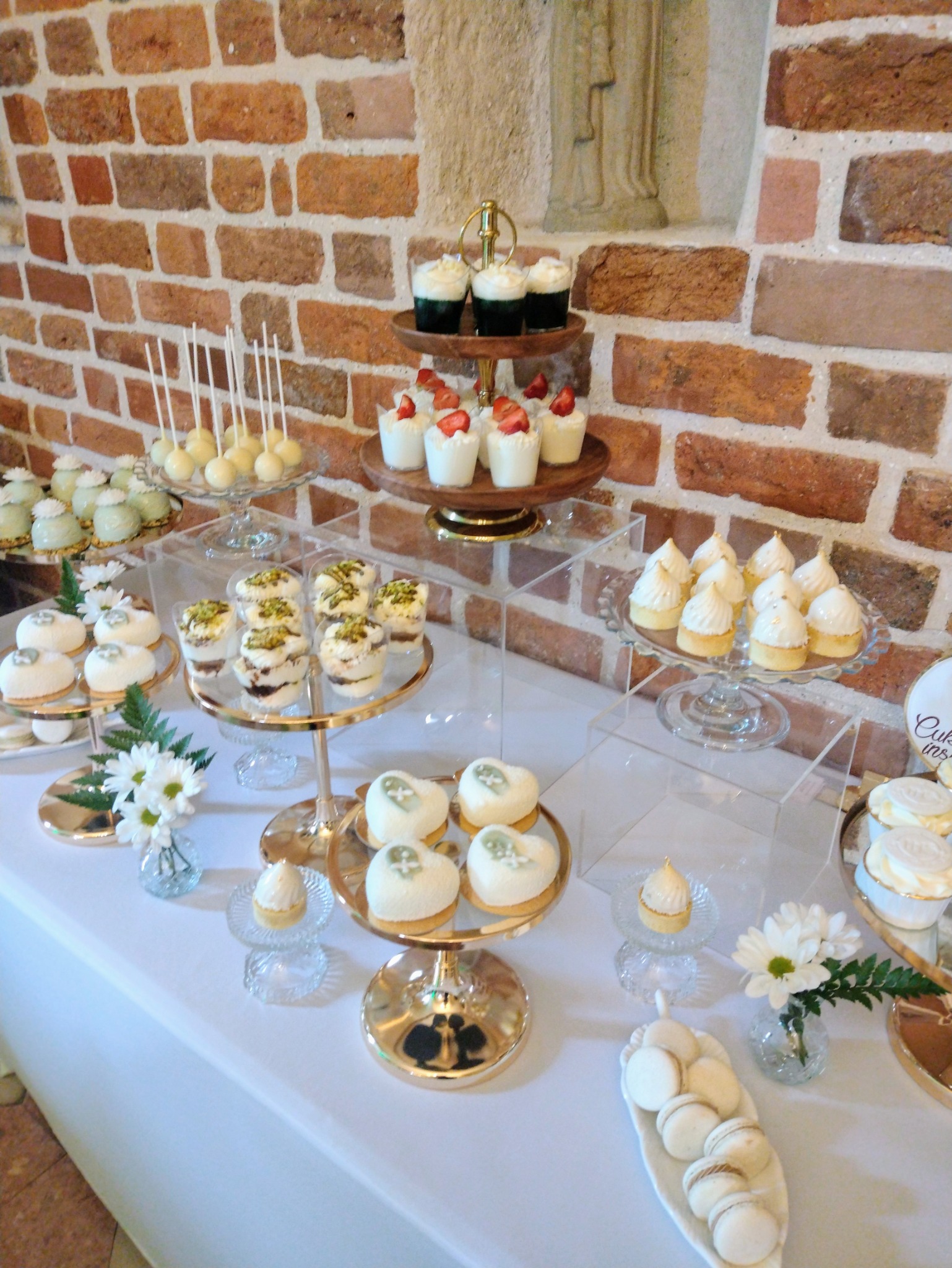 Elegancki stół z deserami: babeczki z truskawkami, cake pops, makaroniki i mini torciki w kształcie serc, ozdobione świeżymi kwiatami na tle ceglanej ściany.