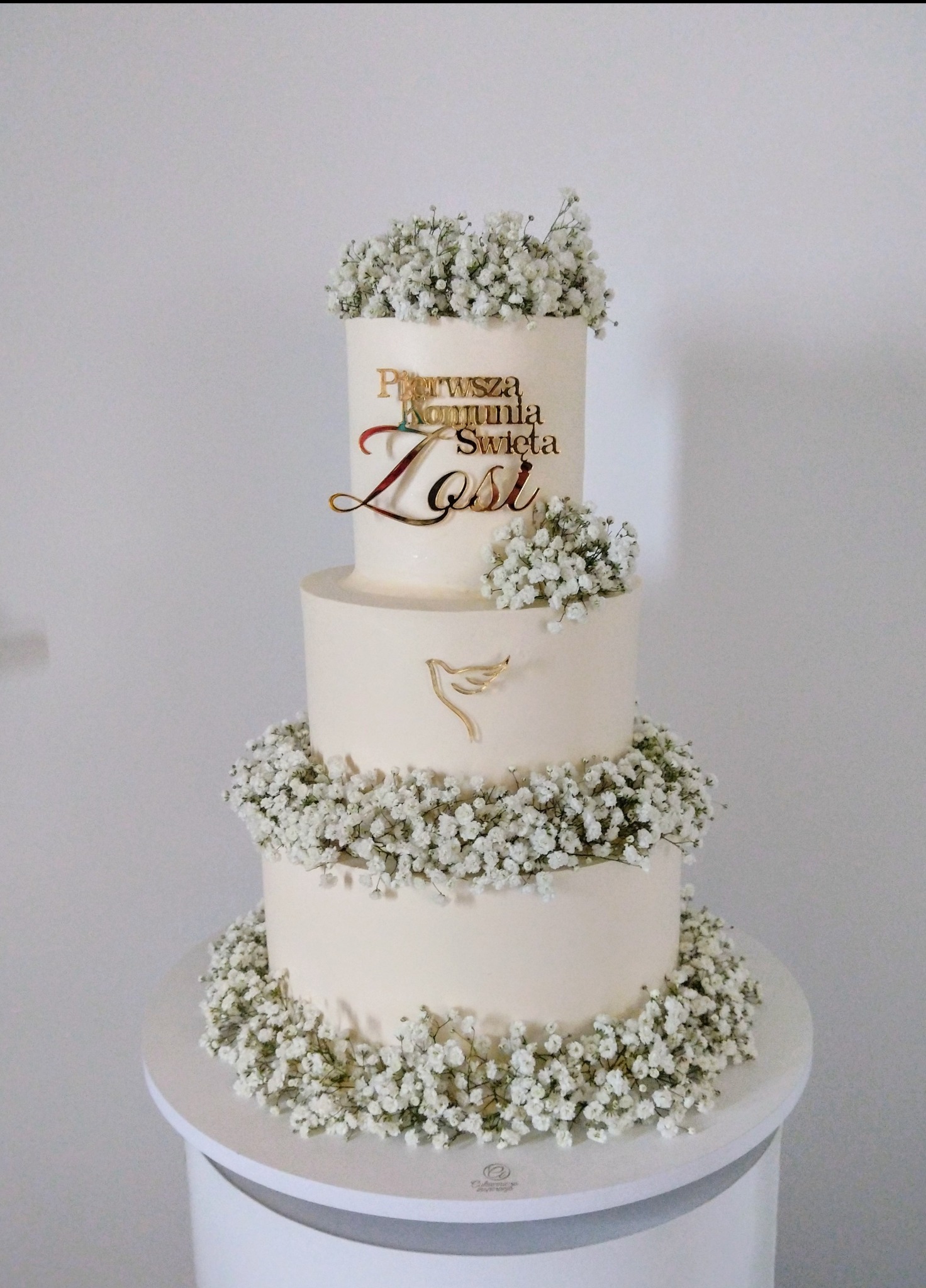 Elegancki, trzypiętrowy tort komunijny w kremowej tonacji, zdobiony białymi kwiatami gipsówki i złotym napisem 'Pierwsza Komunia Święta Zosi'.