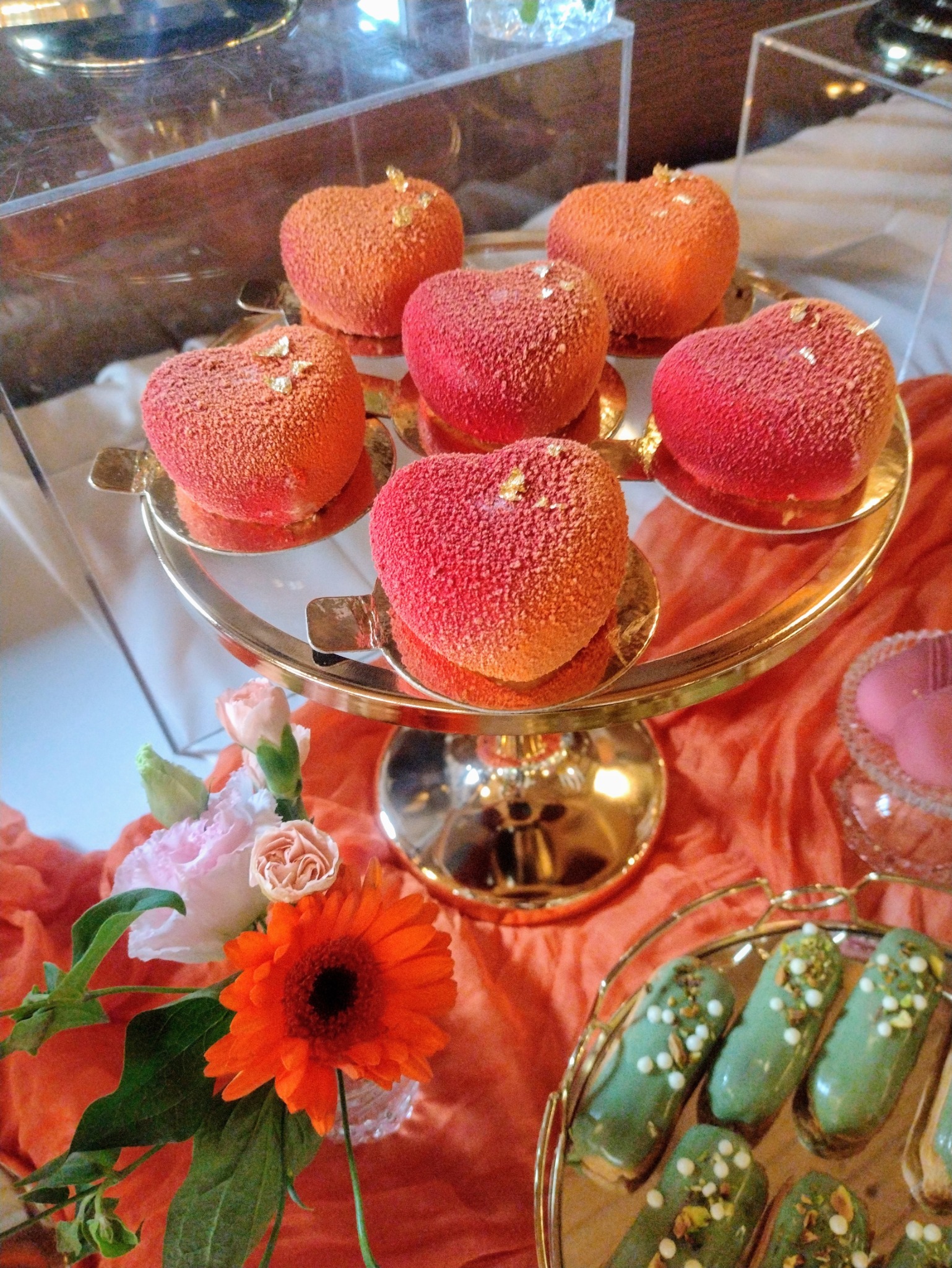 Dekoracyjne ciastka w kształcie serc na złotej paterze, obok eklerki z pistacjami i kwiaty. Elegancka aranżacja słodkiego stołu.