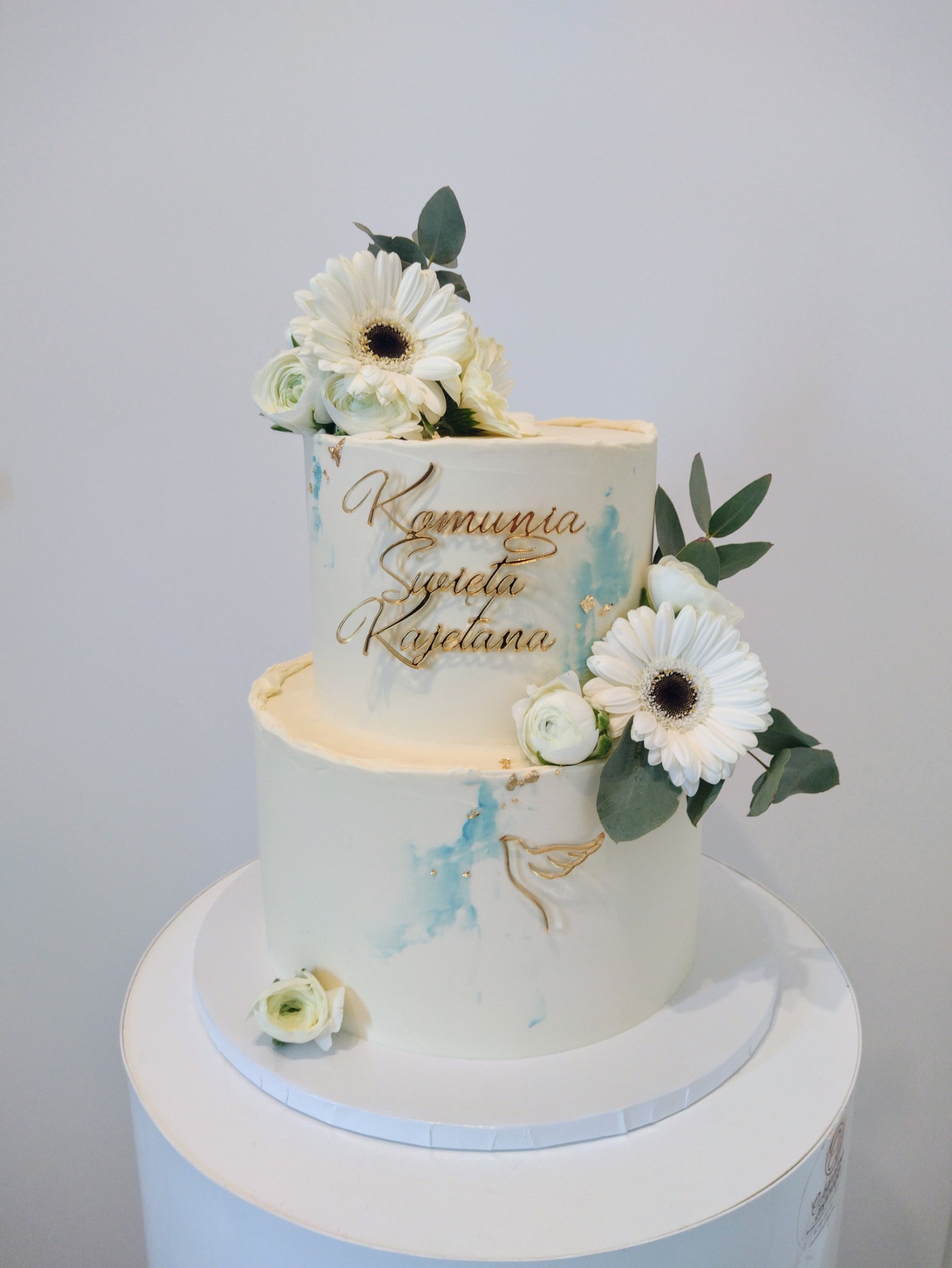 Dwupoziomowy tort na komunię z białym kremem, błękitnymi akcentami, złotym napisem i kwiatami na białym podeście.