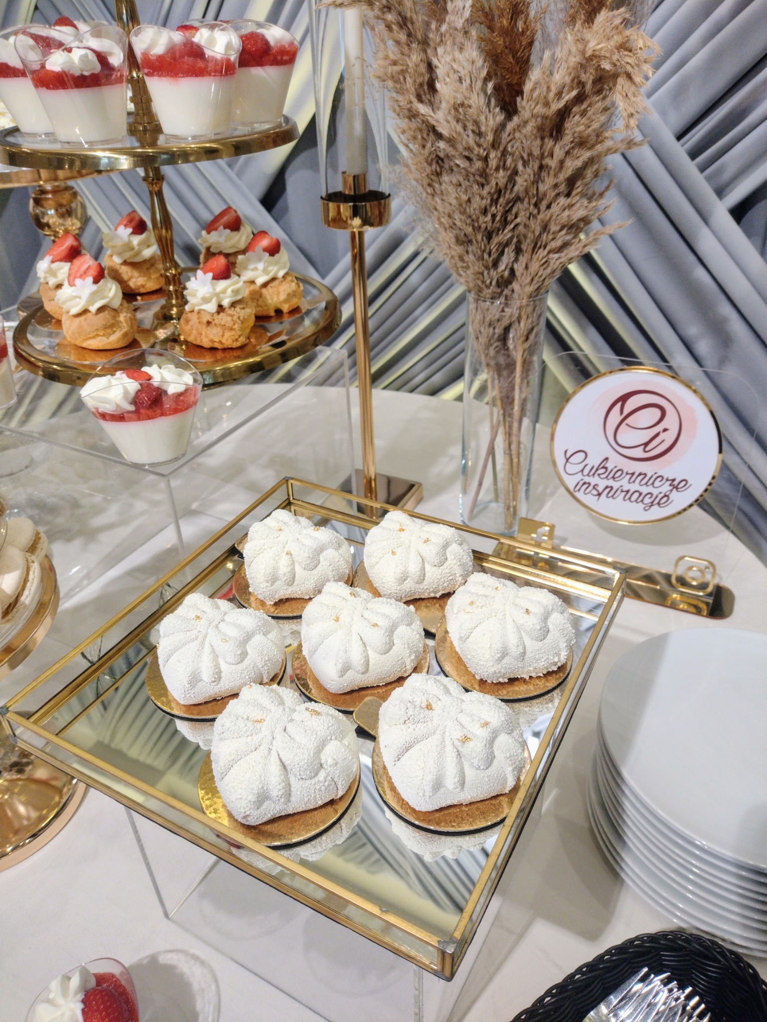 Elegancki bufet słodkości: białe desery na złotych paterach, panna cotta z truskawkami i dekoracje z traw pampasowych. Logo 'Cukiernicze Inspiracje' w tle.