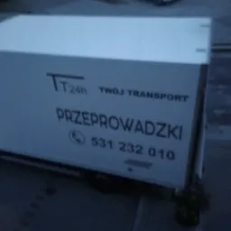 Biała furgonetka z napisem 'TT 24h Twój Transport' i numerem telefonu, zaparkowana na parkingu, widok z góry.