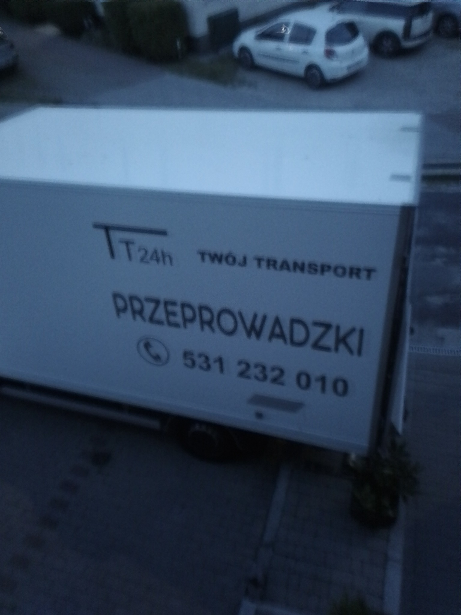 Biała furgonetka z napisem 'TT 24h Twój Transport' i numerem telefonu, zaparkowana na parkingu, widok z góry.