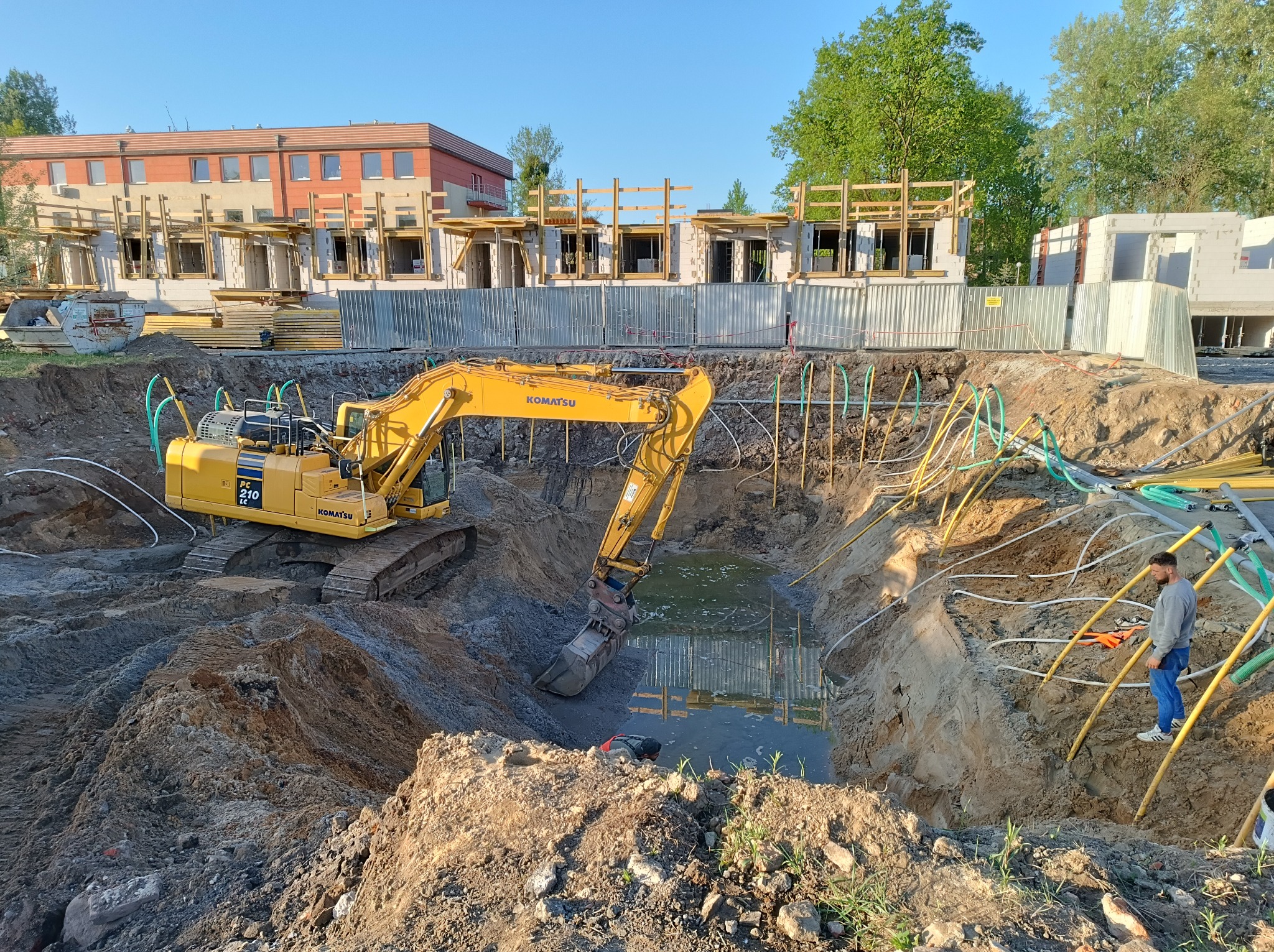 Żółta koparka Komatsu PC210 LC w wykopie wypełnionym wodą, obok widoczny mężczyzna w jeansach, w tle budynek w stanie surowym otwartym i liczne rury drenażowe.