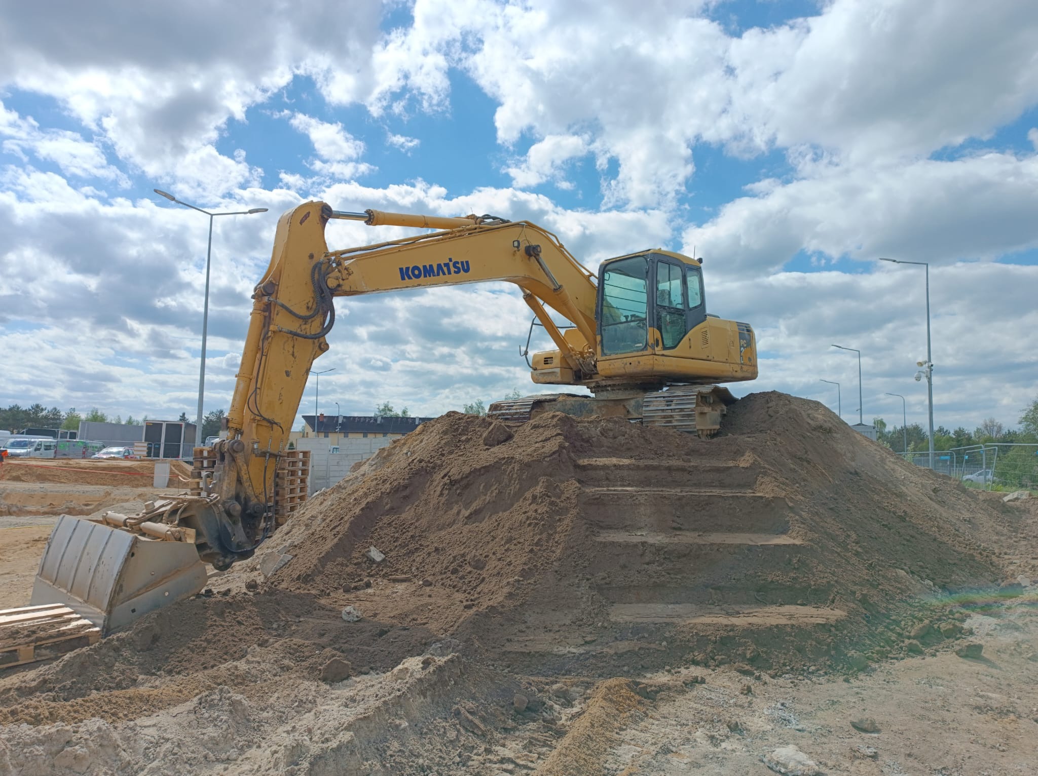 Żółta koparka Komatsu stoi na szczycie usypanej ziemi, tworząc schodkową strukturę, z paletami w pobliżu i błękitnym niebem z chmurami w tle.