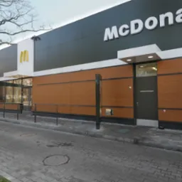 Nowoczesna elewacja budynku restauracji z panelami w kolorze drewna i szarymi elementami, widoczne logo i otoczenie z kostki brukowej.