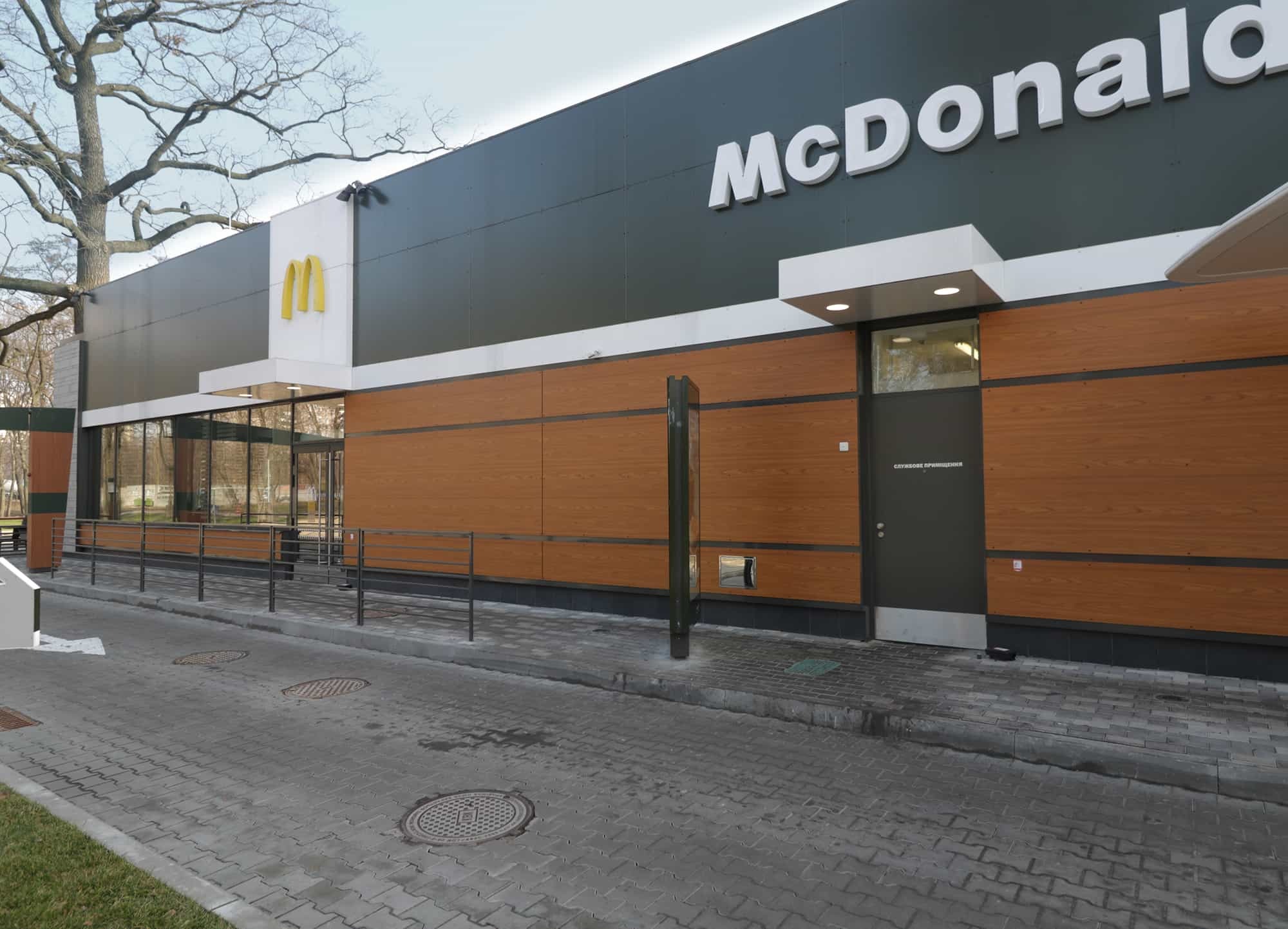 Nowoczesna elewacja budynku restauracji z panelami w kolorze drewna i szarymi elementami, widoczne logo i otoczenie z kostki brukowej.