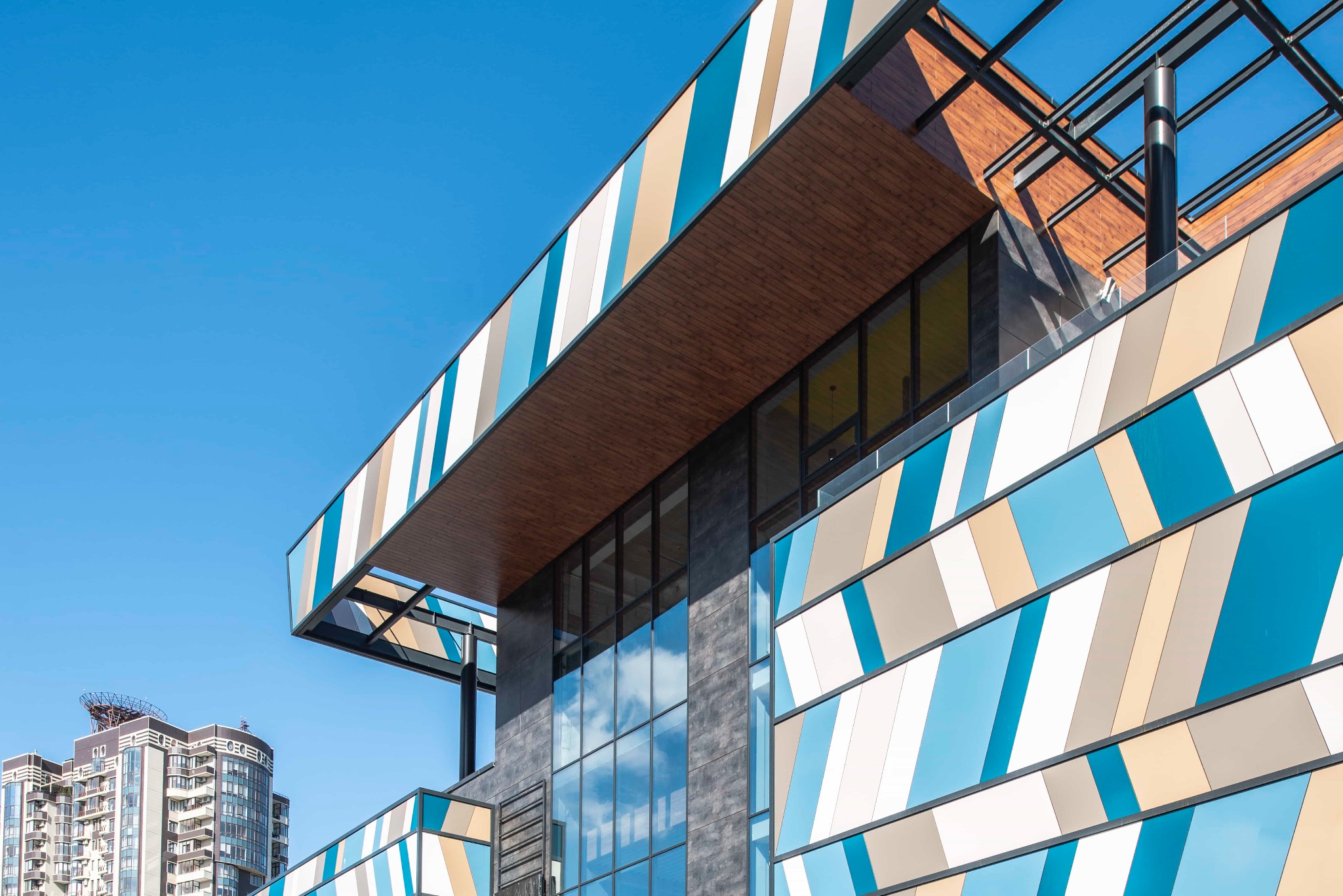 Nowoczesna elewacja budynku CH Novus z panelami w kolorach błękitu, beżu i bieli, tworzącymi geometryczny wzór, widoczna z dołu pod kątem, na tle błękitnego nieba.