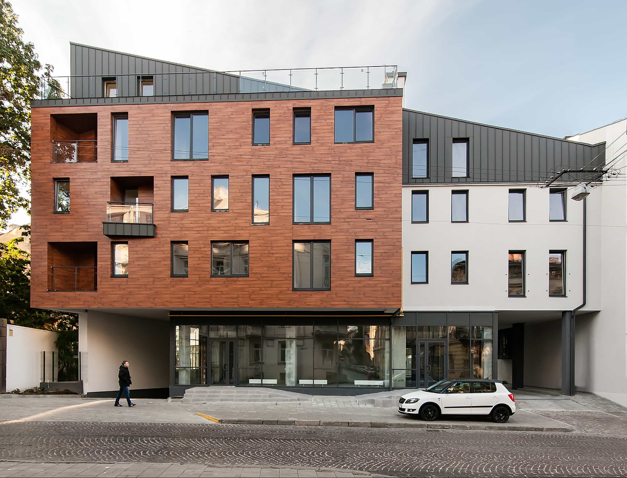 Nowoczesny budynek z elewacją z drewnianych paneli i szarej blachy trapezowej, widok z poziomu ulicy, biały samochód zaparkowany przed wejściem.