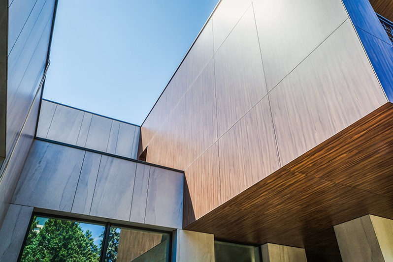 Nowoczesna elewacja budynku z paneli imitujących drewno i marmur, widziana z dołu pod kątem, z fragmentem okna i niebem w tle.