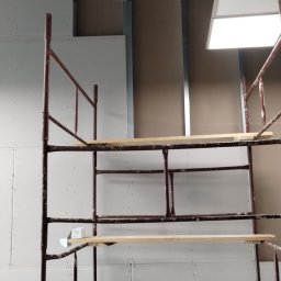 Dominik Róg - Montaż płyt karton gips na metalowym stelażu, widoczne rusztowanie malarskie. Ściana z zamontowanymi płytami, widoczne łączenia i śruby. Oświetlenie sufitowe.