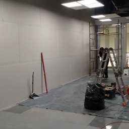 Dominik Róg - Montaż płyt karton-gips na ścianie w biurze. Widoczny pracownik na rusztowaniu, narzędzia, poziomica i posprzątana podłoga. Oświetlenie sufitowe.