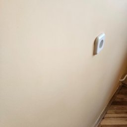 Dominik Róg - Ściana w odcieniach beżu z zamontowanym gniazdkiem elektrycznym. Widoczna drewniana podłoga i listwa przypodłogowa. Delikatne oświetlenie.