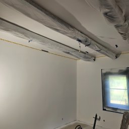 Dominik Róg - Białe ściany po malowaniu natryskowym, zabezpieczone folią sufit i okno, widoczny agregat malarski i drabina. Świeżo pomalowane wnętrze.