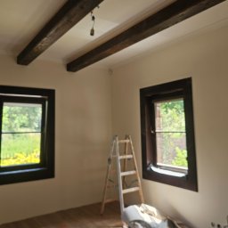 Dominik Róg - Pokój po malowaniu ścian na beżowo z drewnianymi belkami stropowymi i oknami w ciemnych ramach. Widoczna drabina i zabezpieczenia podłogi.