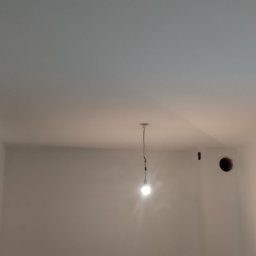 Dominik Róg - Wnętrze pokoju po szpachlowaniu ścian, widoczne gładkie, białe ściany, lampa wisząca i halogeny oświetlające. Przy podłodze widoczne gniazdka elektryczne.