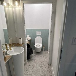 Dominik Róg - Nowoczesna łazienka z umywalką na postumencie, płytkami w odcieniach szarości i błękitu. Widok z korytarza, oświetlenie punktowe i minimalistyczny design.