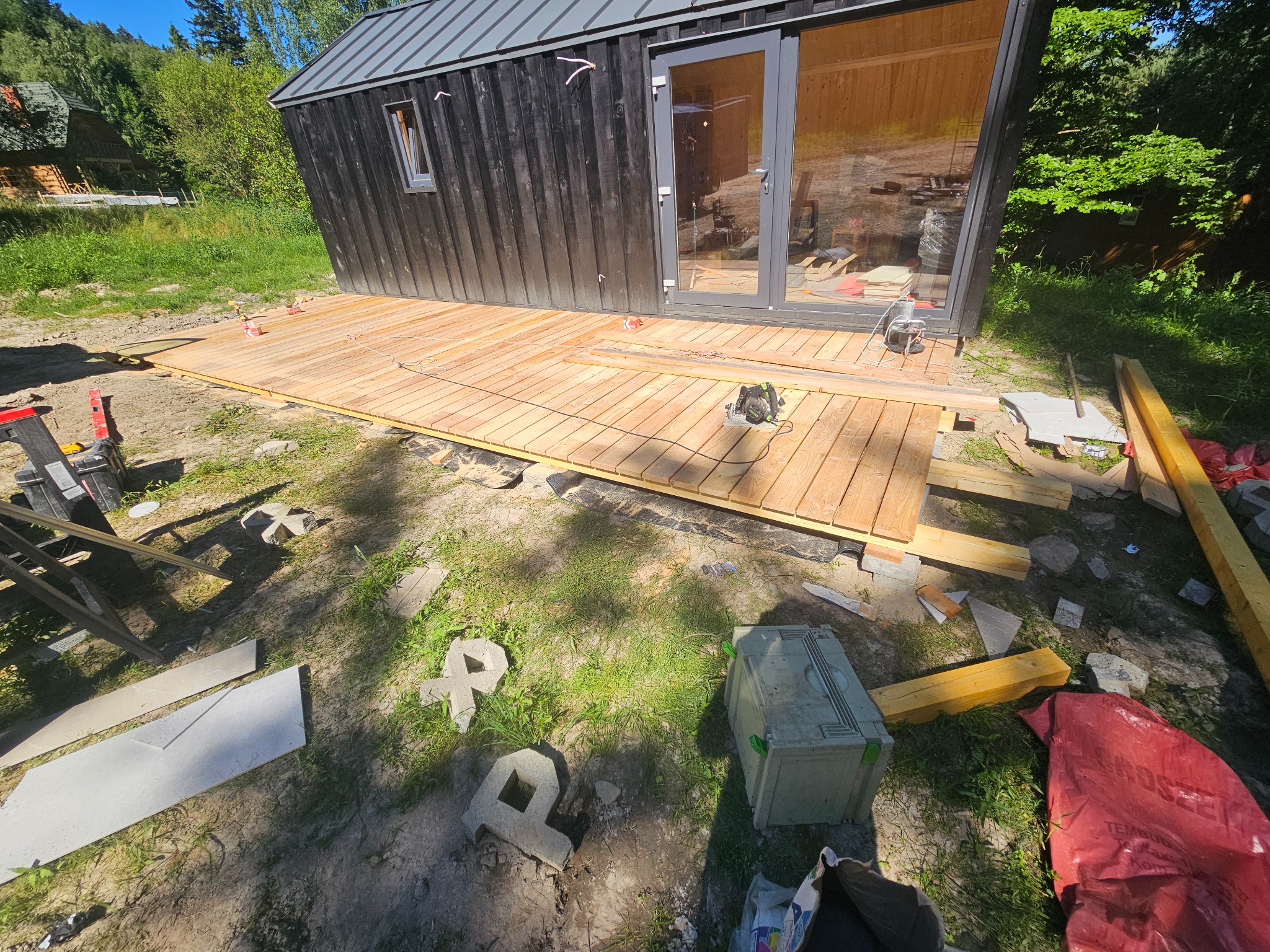 Budowa drewnianego tarasu przy czarnym domku letniskowym. Widoczne narzędzia, deski i elementy konstrukcyjne. Prace w toku, słoneczny dzień.