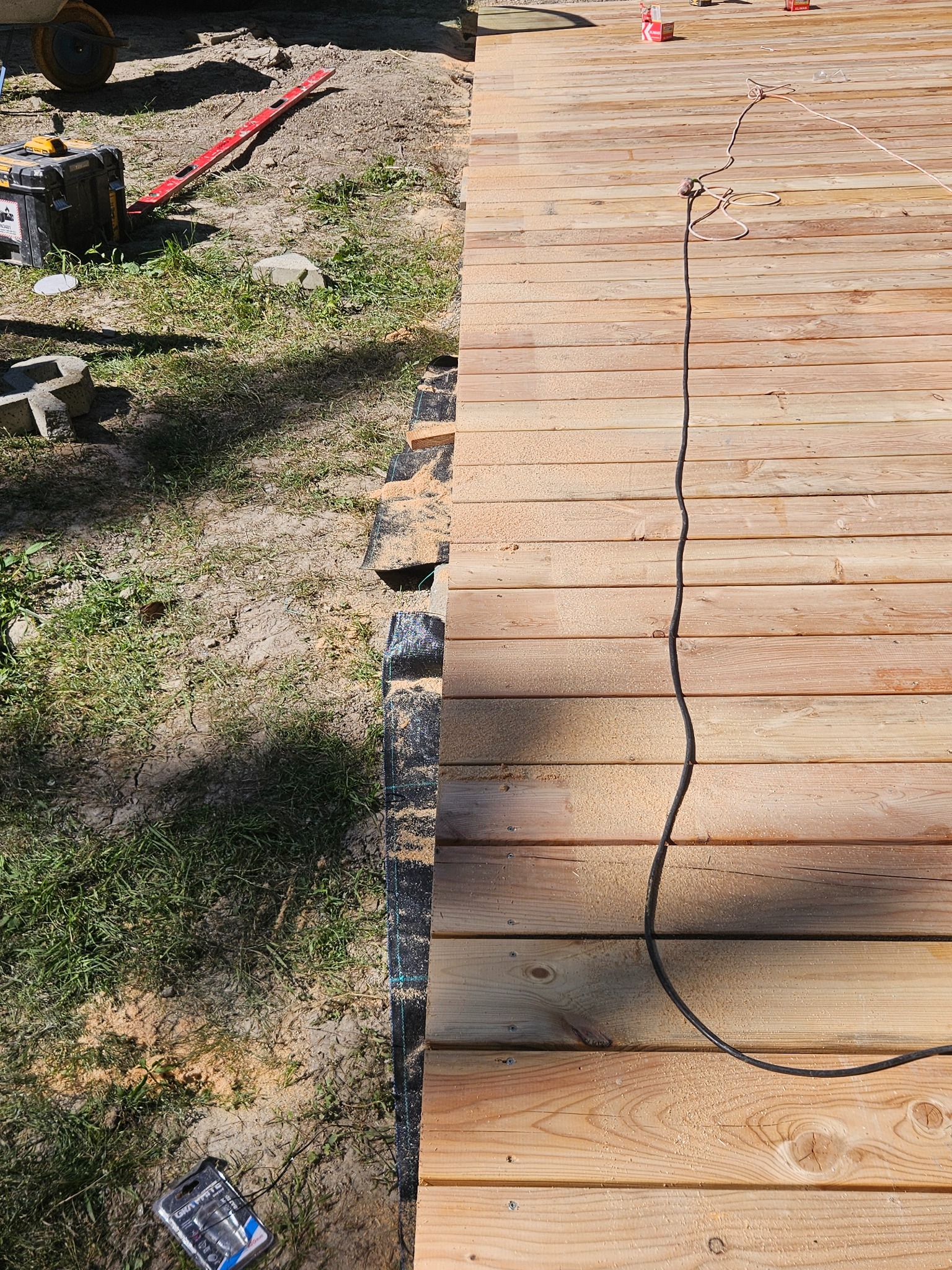 Fragment nowej drewnianej podłogi tarasu w trakcie budowy, widoczne deski, narzędzia i kabel. Ujęcie z góry, oświetlenie słoneczne.