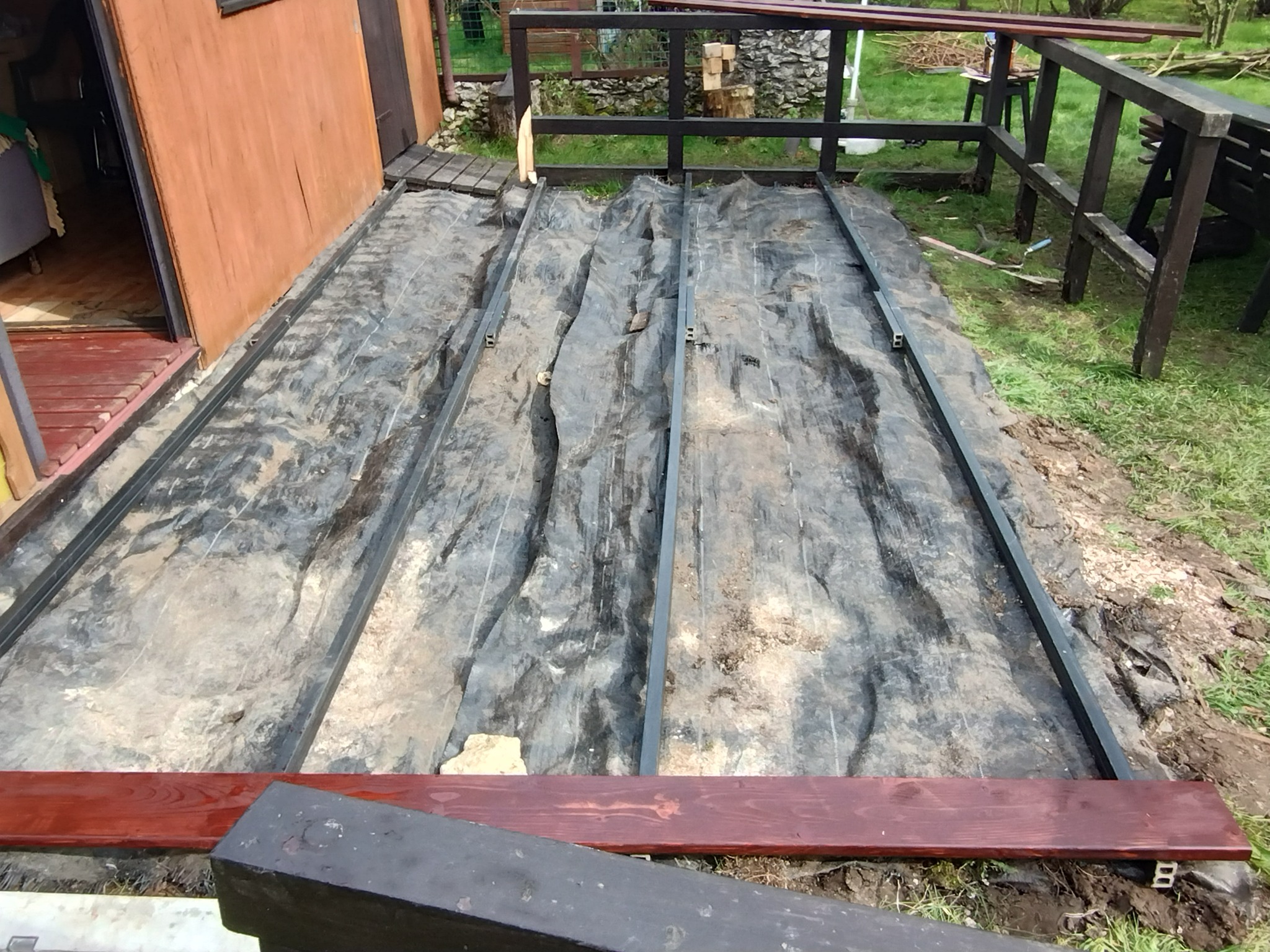 Montaż konstrukcji pod taras: metalowe legary na folii, przygotowane deski w kolorze mahoniu. Widok z góry na plac budowy tarasu przy budynku.