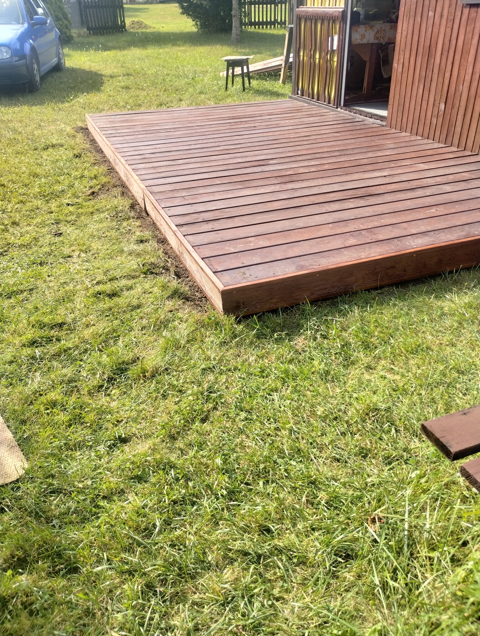 Drewniany taras z desek na trawie, przylegający do budynku z drewnianymi elementami. Widoczny fragment niebieskiego samochodu i stołek w tle.