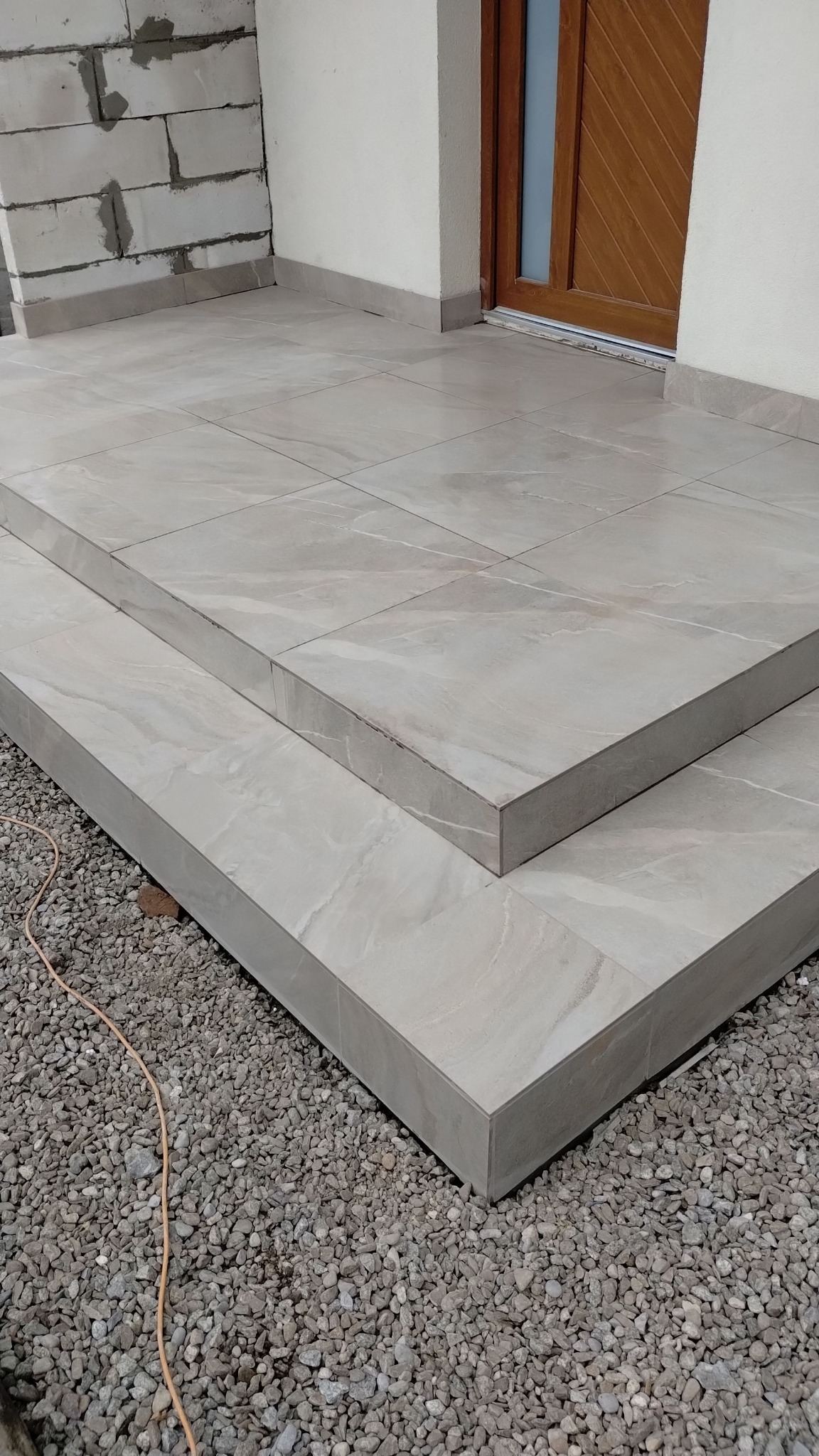 Wykończone płytkami schody wejściowe z szarymi fugami, prowadzące do drewnianych drzwi. Tło stanowią bloczki betonowe i szare kamienie.