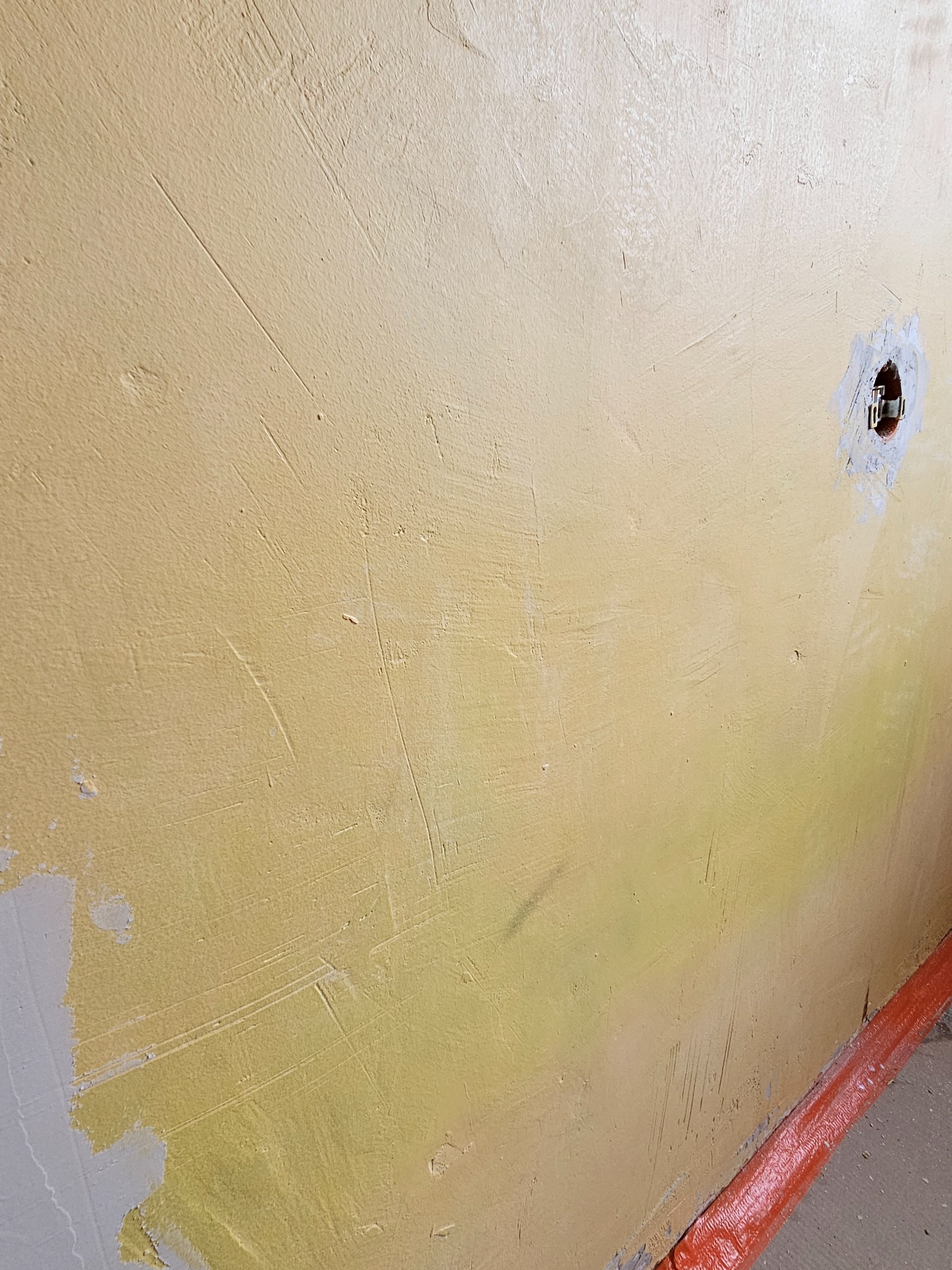 Żółta ściana w trakcie malowania, widoczne ślady pędzla i niedomalowane fragmenty. Otwór na gniazdko elektryczne przygotowany do wykończenia. Listwa przypodłogowa zabezpieczona.