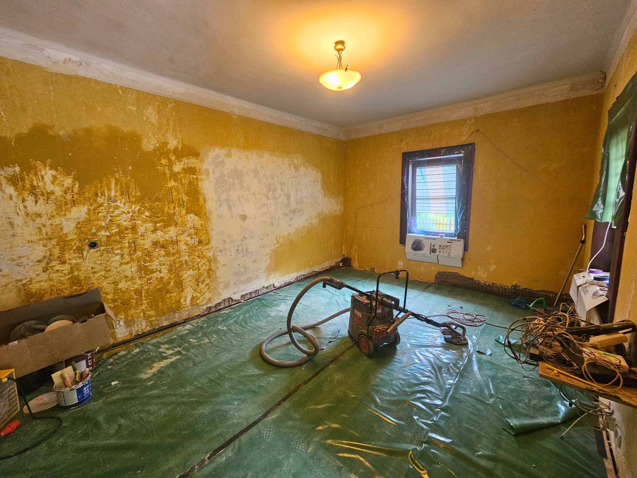 Pokój w trakcie przygotowania do remontu: ściany częściowo zdarte z farby, podłoga zabezpieczona folią, widoczna szlifierka do gładzi i narzędzia malarskie.